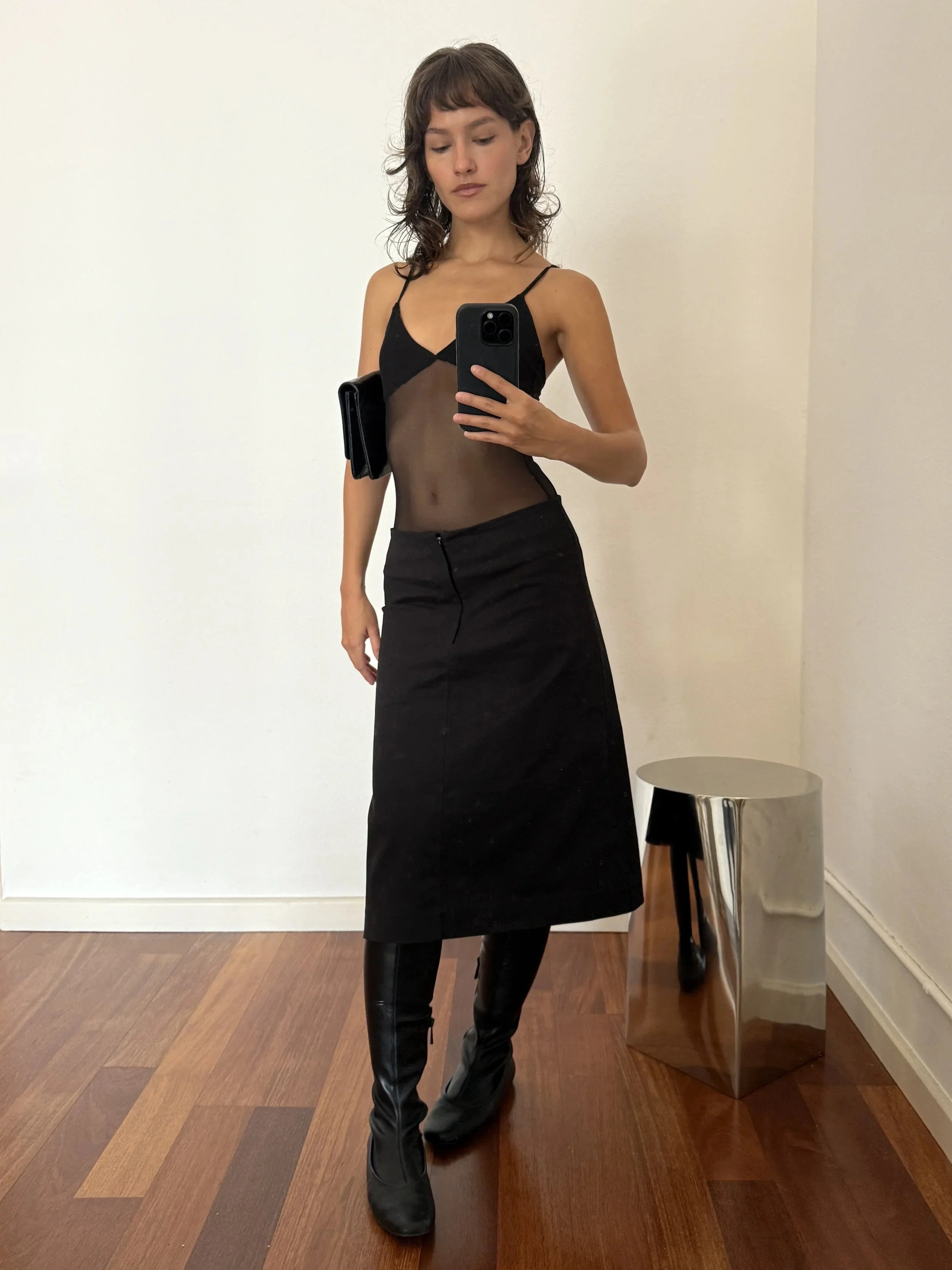 A woman standing indoors taking a mirror selfie. She is wearing a black sheer top with spaghetti straps, a black skirt, and black leather pants. She is holding a phone in her right hand and has a black clutch purse under her right arm. There is a sma