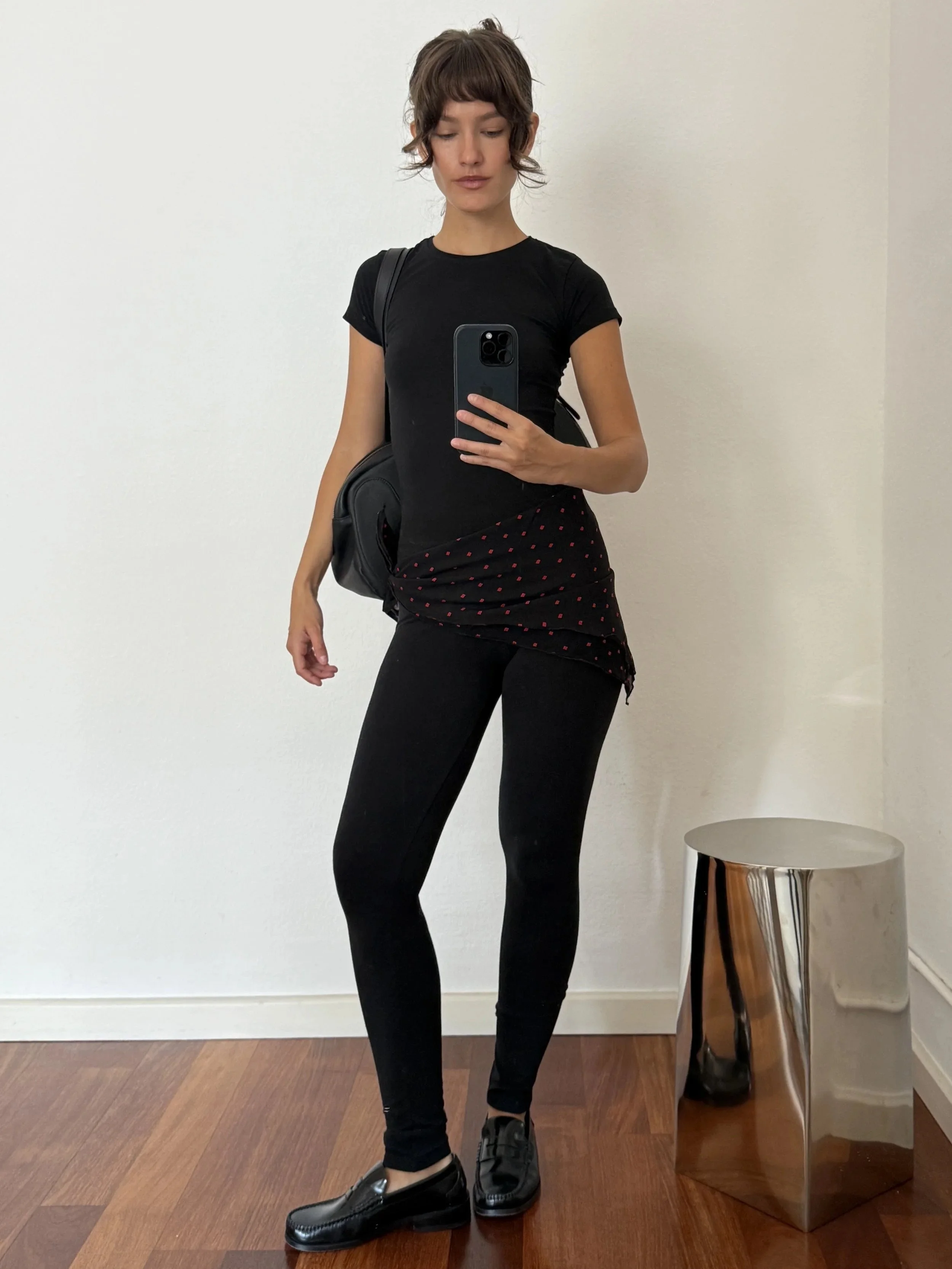Woman with short brown hair taking a mirror selfie in a plain room, dressed in black pants, black shirt, and black shoes. She carries a black backpack and stands near side table with a metallic finish.