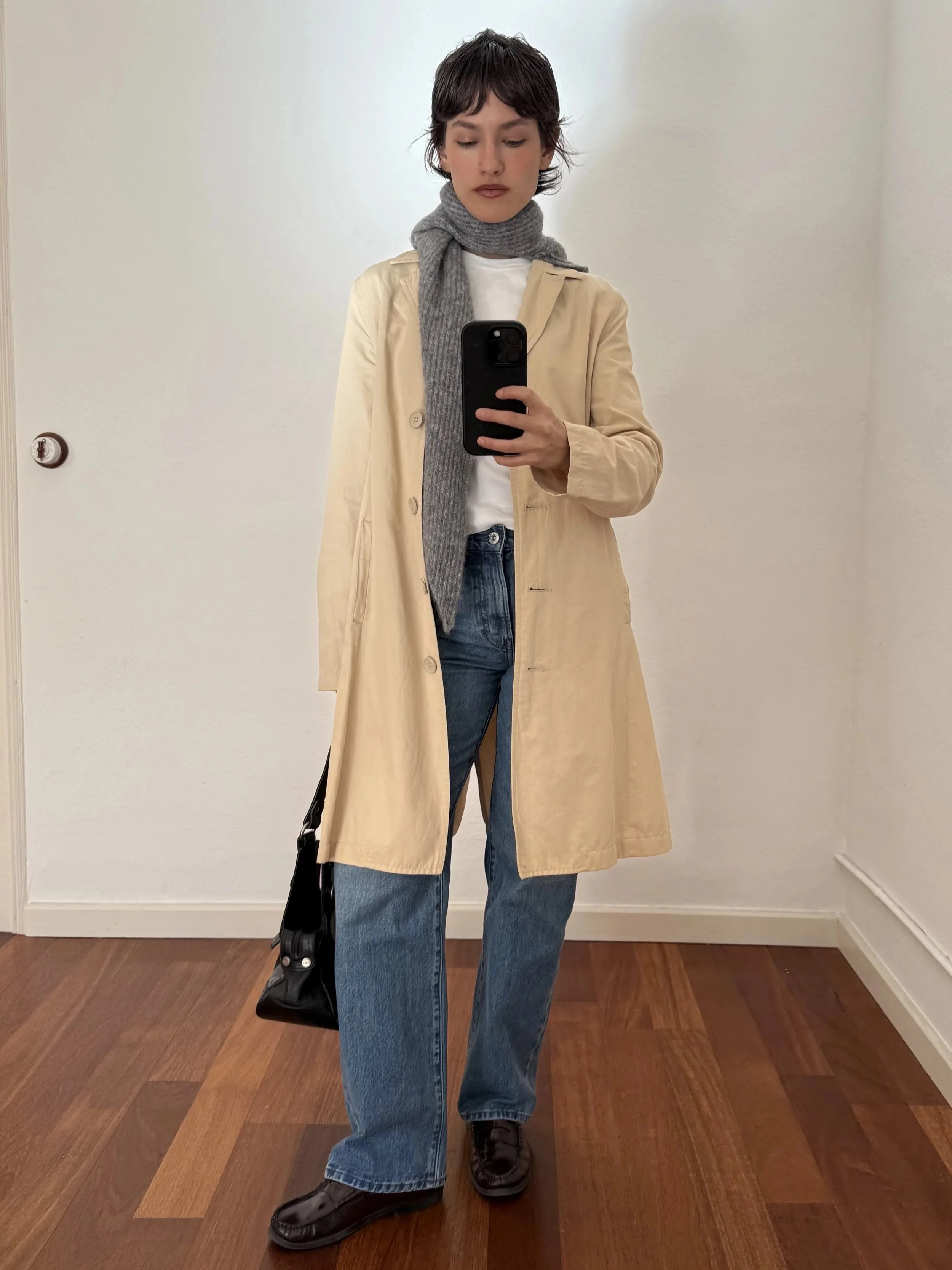 Person taking a mirror selfie, wearing a beige trench coat, gray scarf, white T-shirt, blue jeans, and black boots, with a black bag hanging from one shoulder, standing on a wooden floor against plain white walls.
