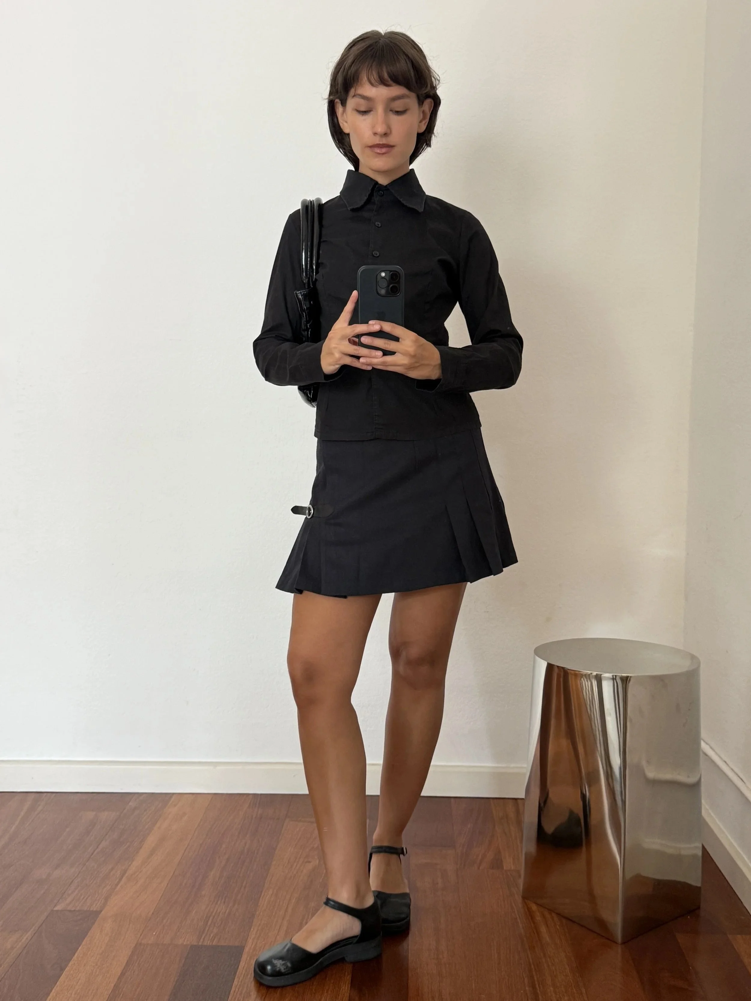A woman taking a mirror selfie, wearing a black long-sleeve button-up shirt, black pleated skirt, black shoes, and black socks, standing on a wooden floor.