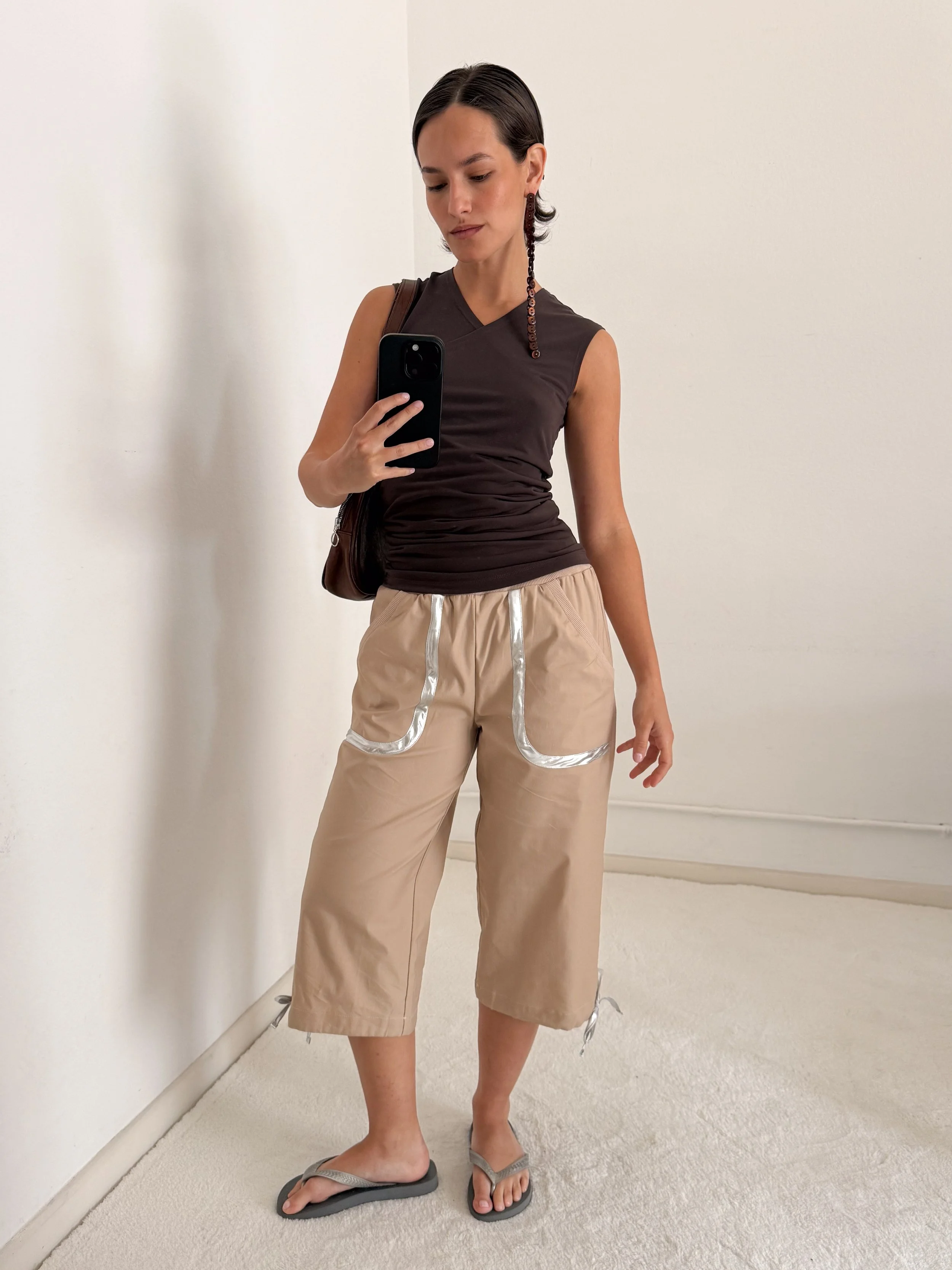 A woman taking a mirror selfie, wearing a dark sleeveless top, beige pants with reflective stripes, flip-flops, and carrying a brown shoulder bag, standing on a beige carpeted floor in front of a plain white wall.