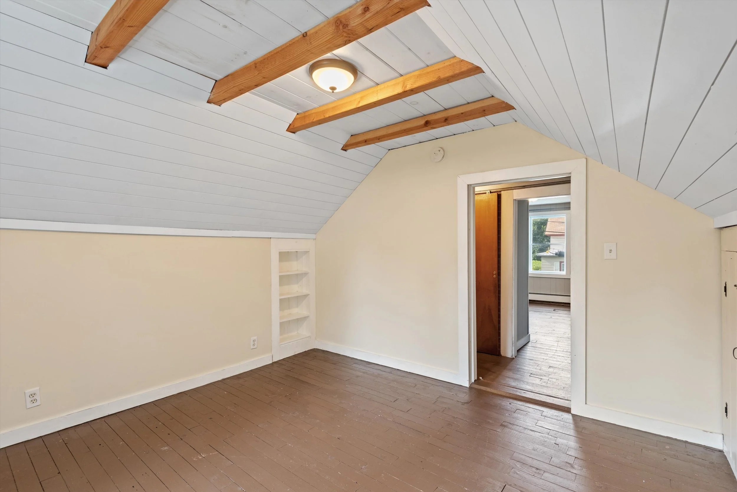 Interior of a small room with a sloped ceiling, white walls, brown wooden floor, and an open doorway leading to a brighter room with a window.