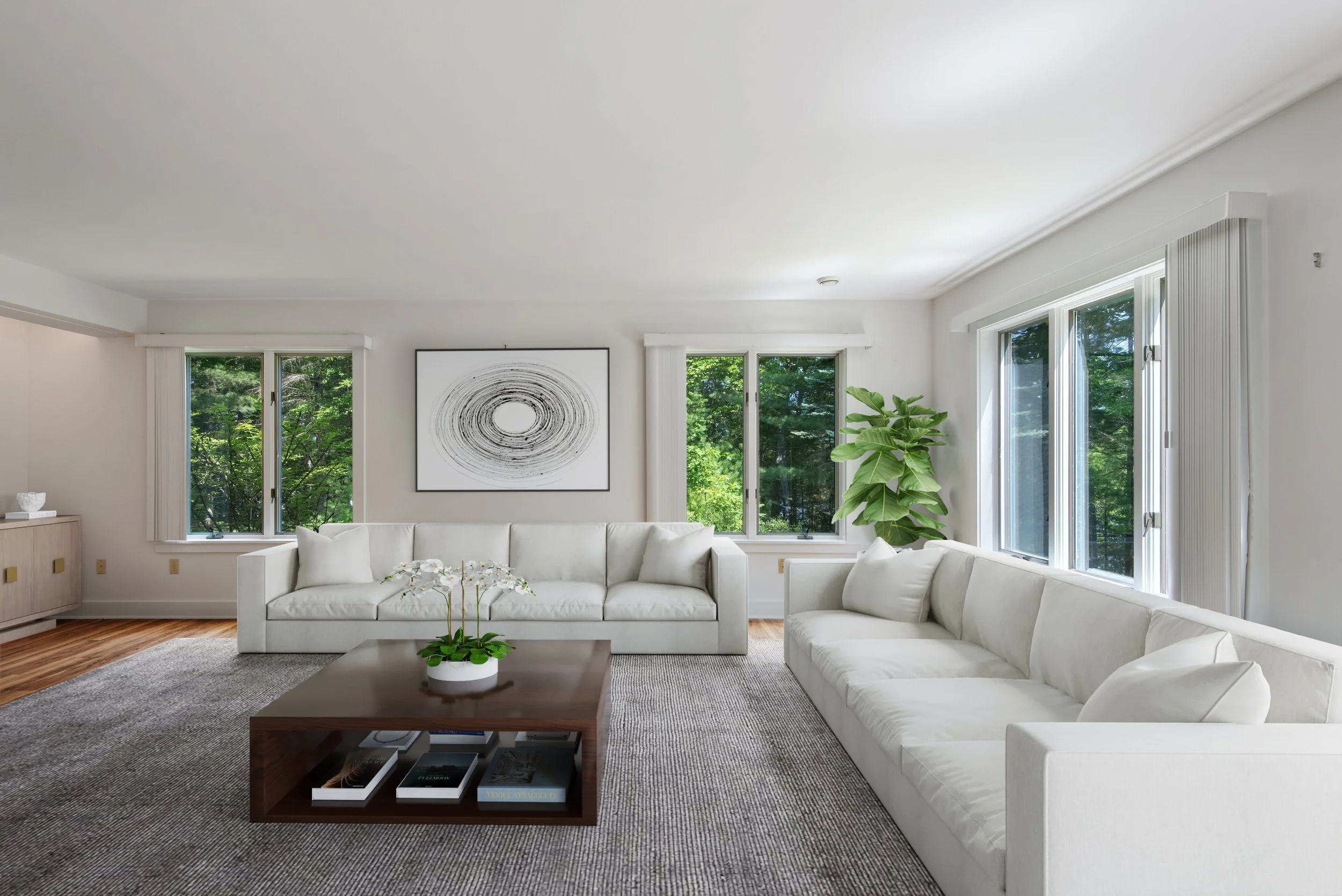 Bright living room with white sofas, wooden coffee table with books and plant, large windows with green trees outside, abstract art on wall, and hardwood floor.