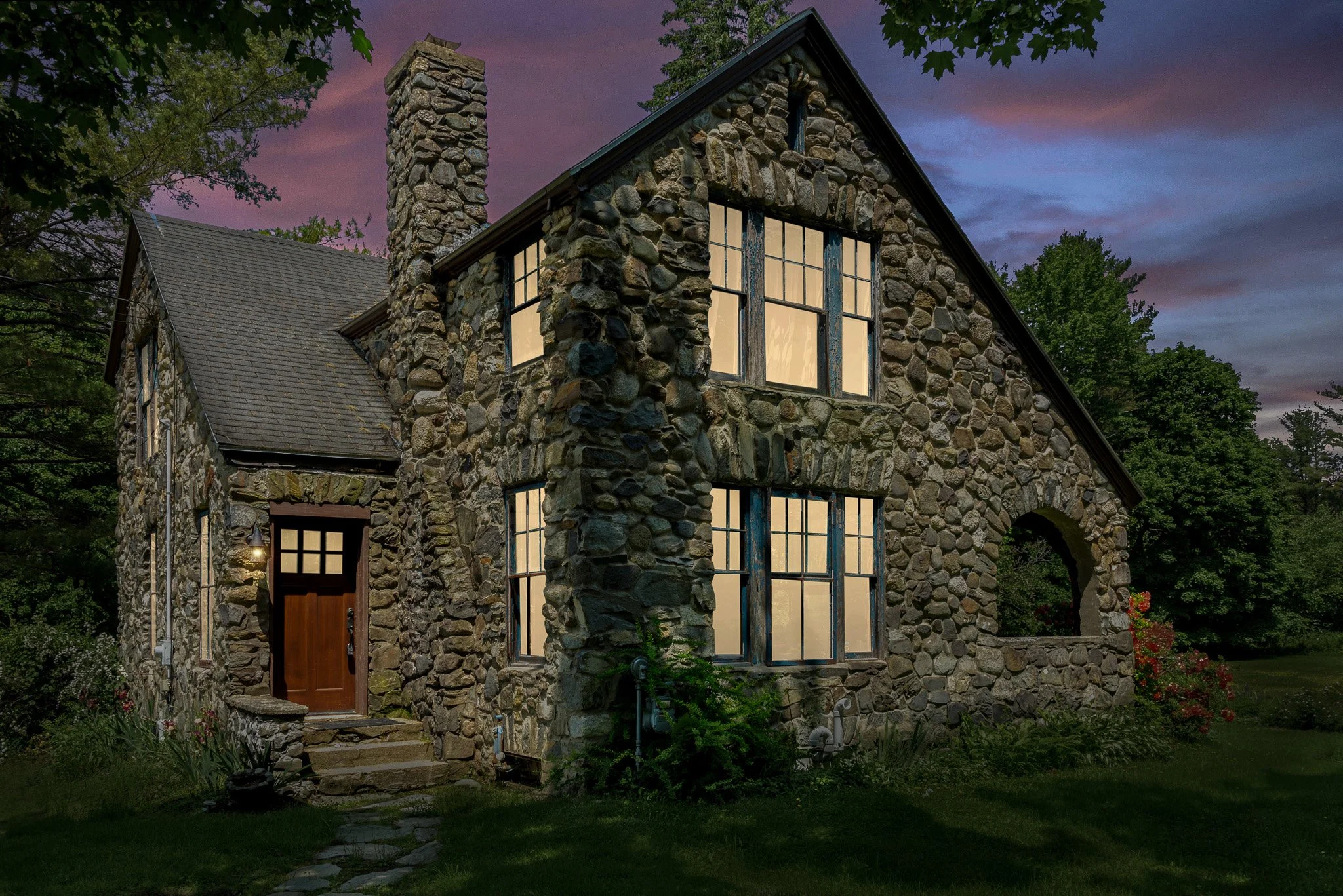 Stone cottage with large windows glowing from inside, set in a lush green yard during dusk, with a cloudy sky and trees in the background.