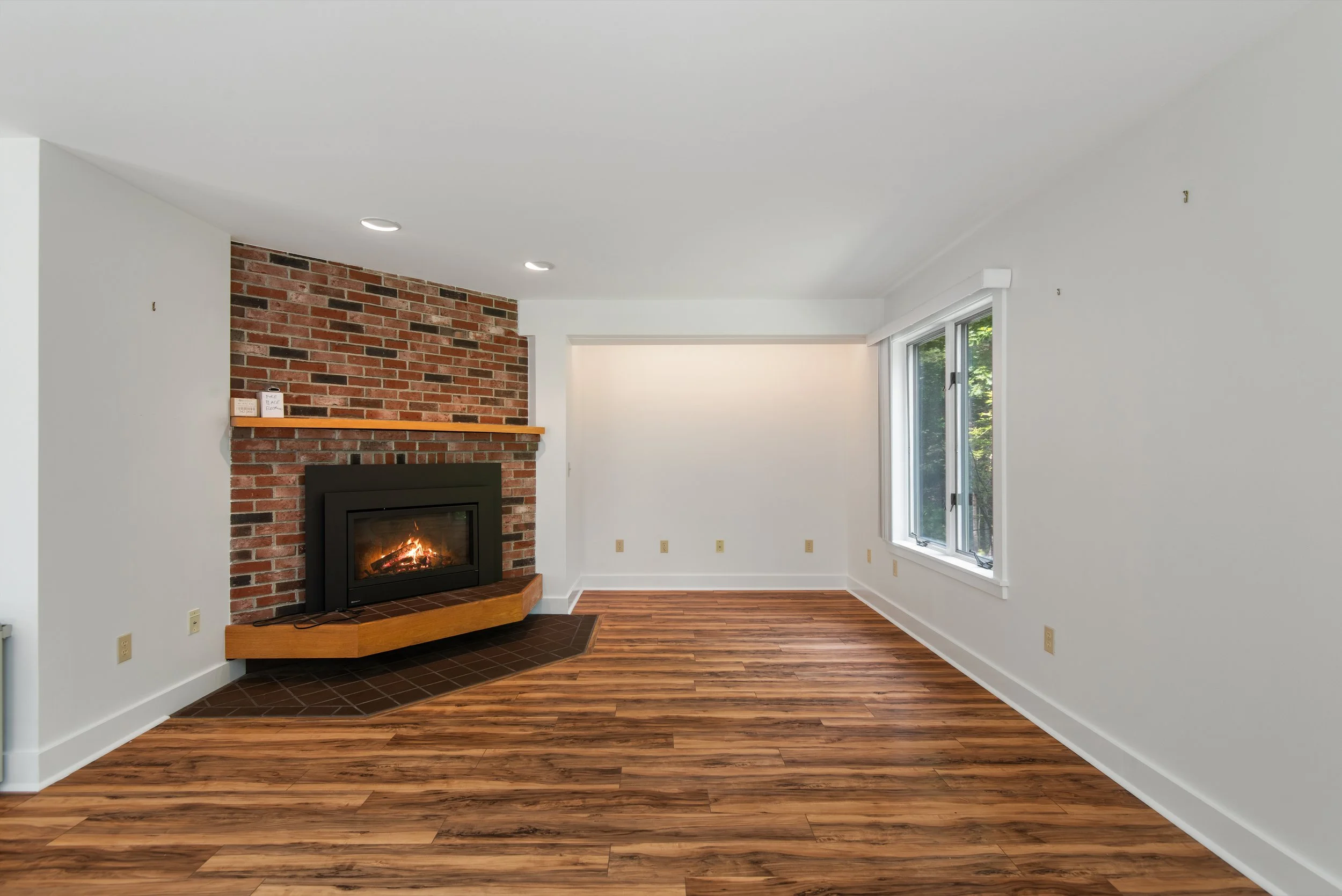 Empty living room with a brick fireplace featuring a wood mantel, hardwood flooring, and large windows showing greenery outside.