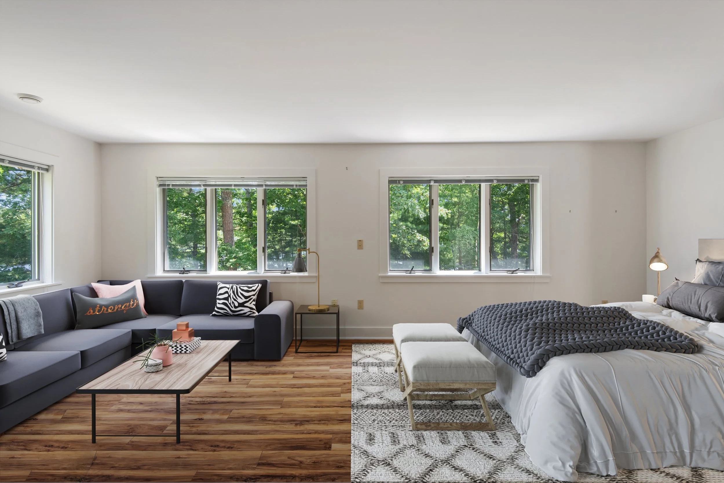 Living room with a gray sectional sofa, pink and black patterned pillows, a wooden coffee table, a side table with a lamp, a bed with dark gray throw, a bench, and large windows showing green trees outside.