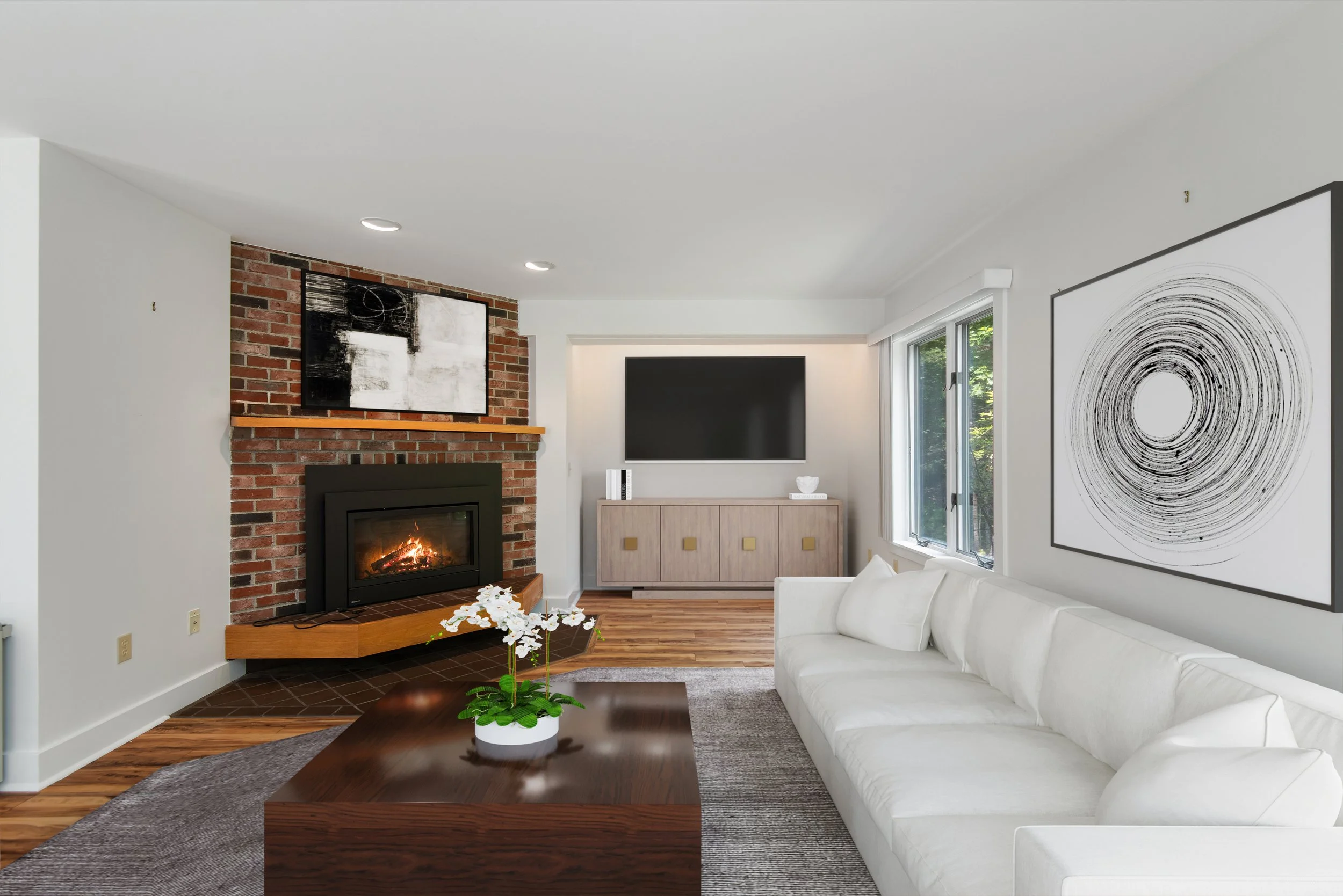 Living room with a white sofa, a wooden coffee table with a potted orchid, a brick fireplace with a wooden mantel, a wall-mounted TV above a light wood cabinet, and modern abstract artwork on the walls near a window.