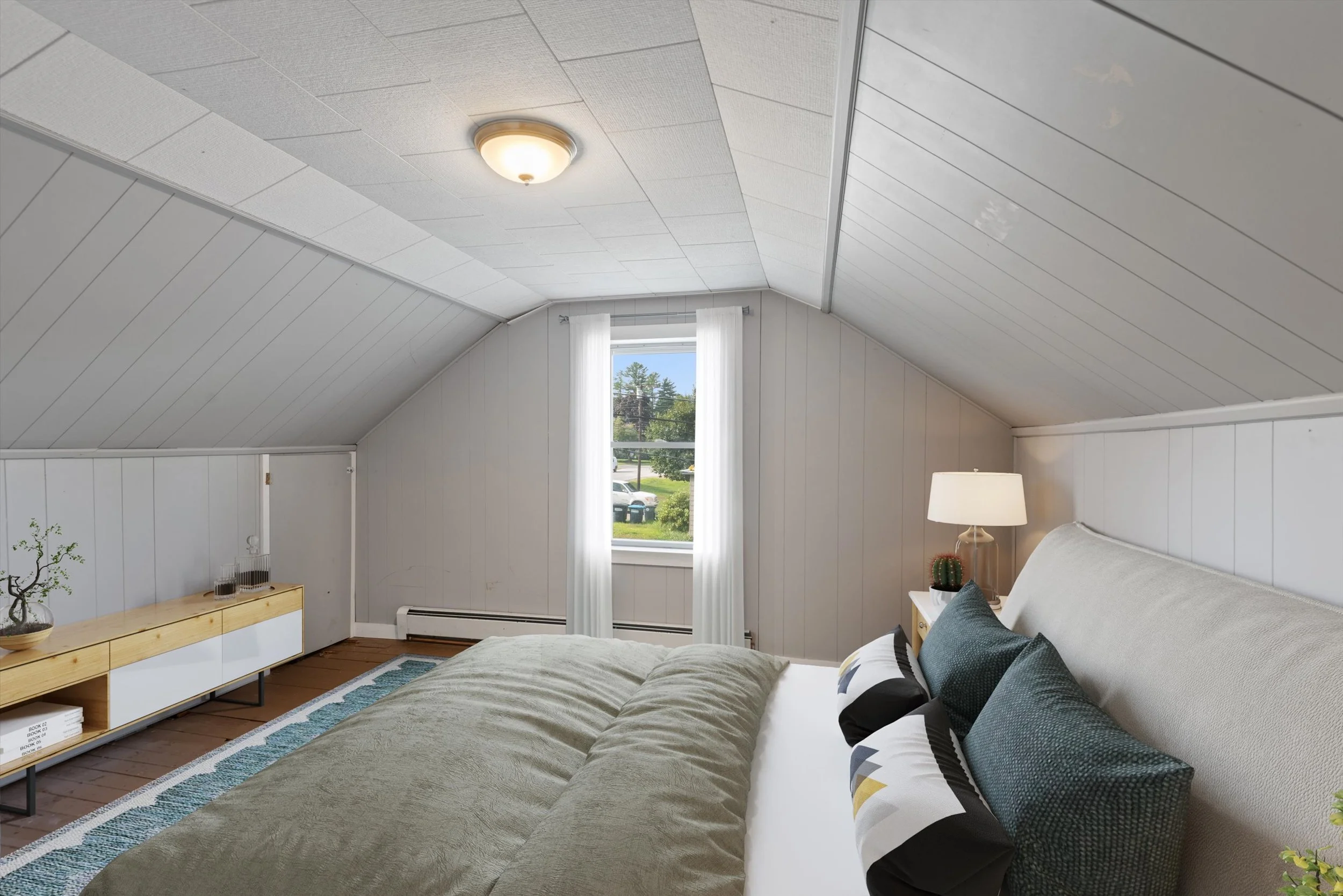 Bedroom with sloped ceiling, white walls, a window with curtains, a bed with pillows, a nightstand with a lamp and cactus, and a wooden console table.