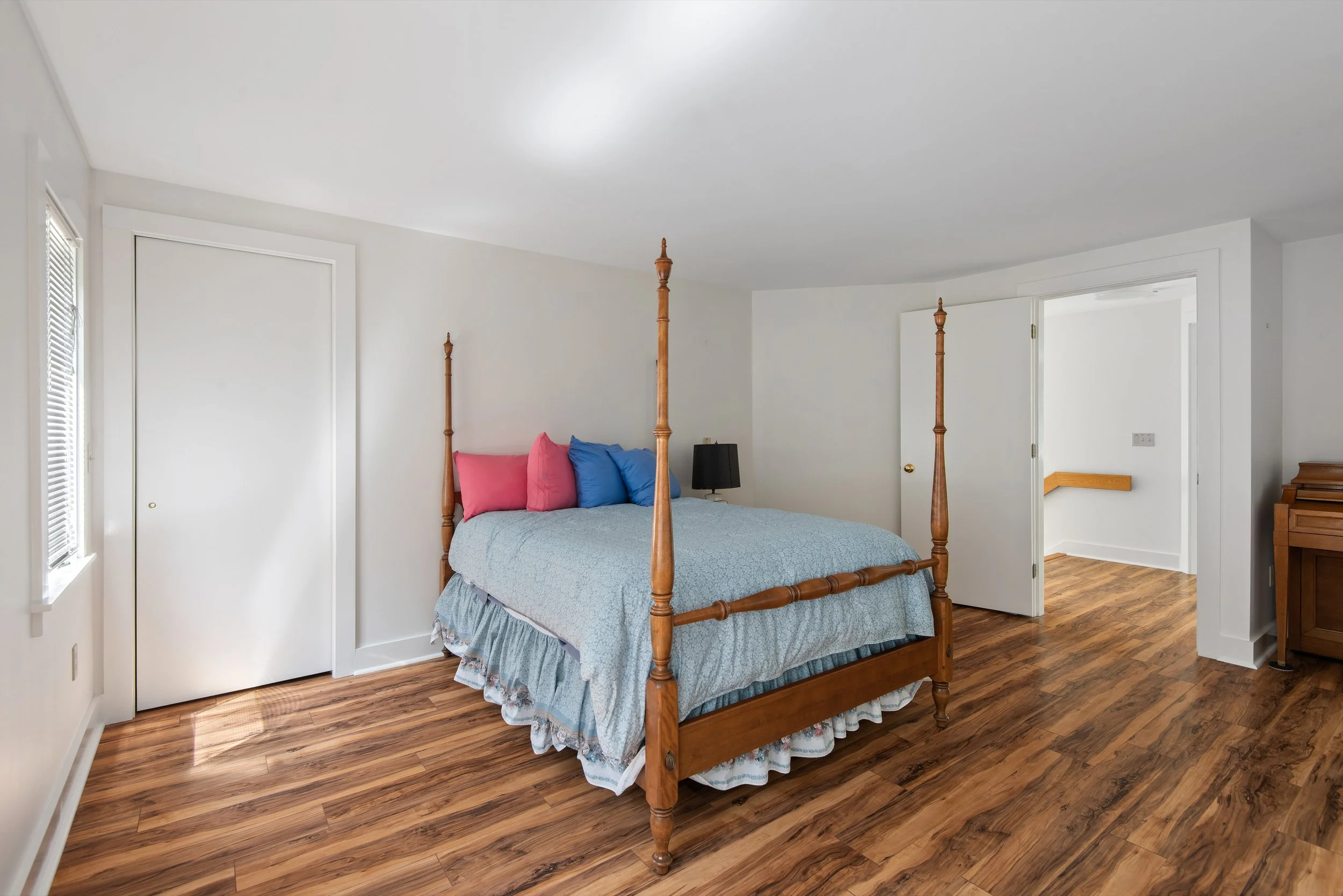 Bedroom with wood floor, white walls, four-poster bed with blue bedding and colorful pillows, black nightstand with lamp, and open door leading to a small room or hallway.
