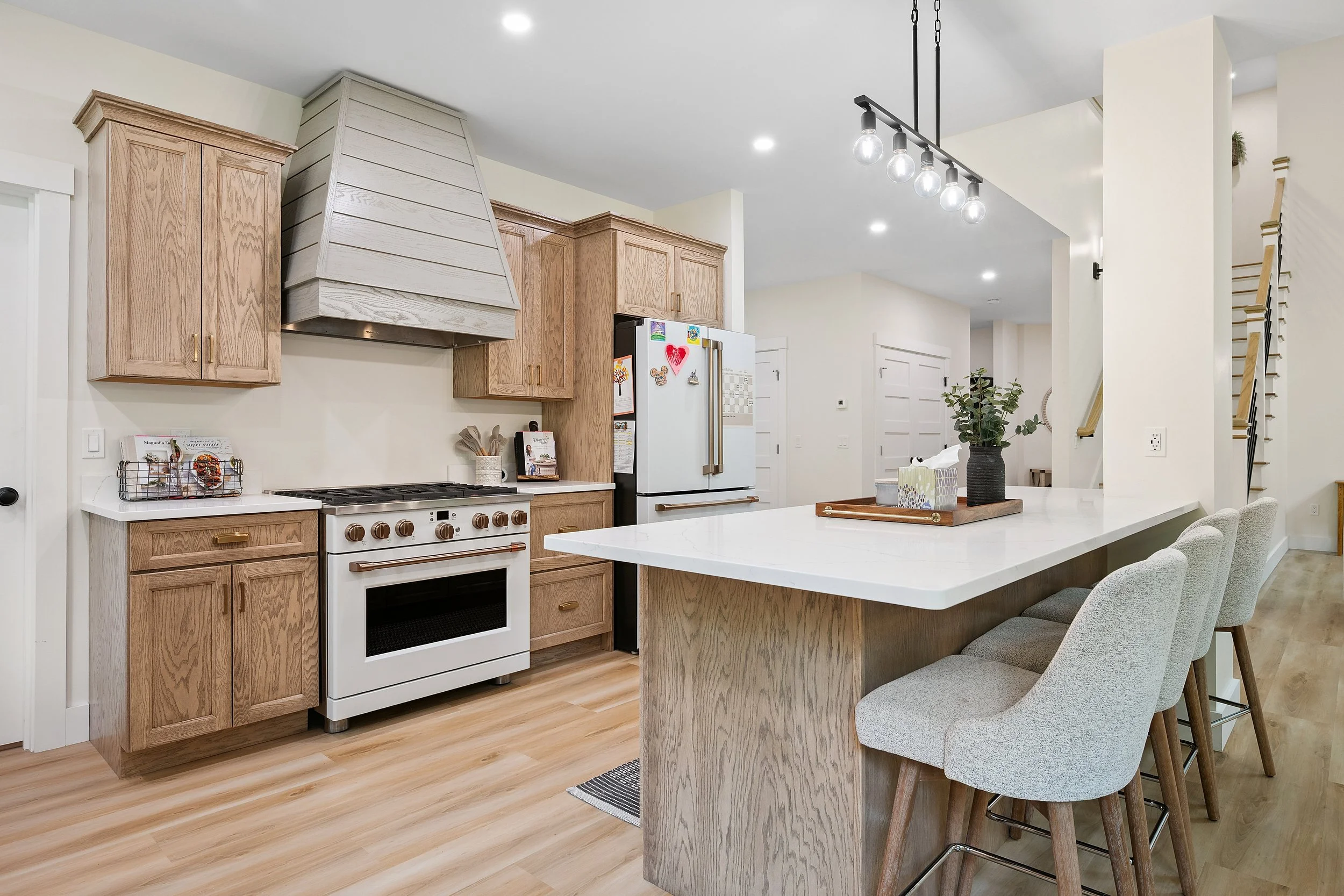 Modern kitchen with wooden cabinets, white countertops, a white refrigerator with magnets, a stove, and bar stools at a breakfast bar, accented by a black hanging light fixture.