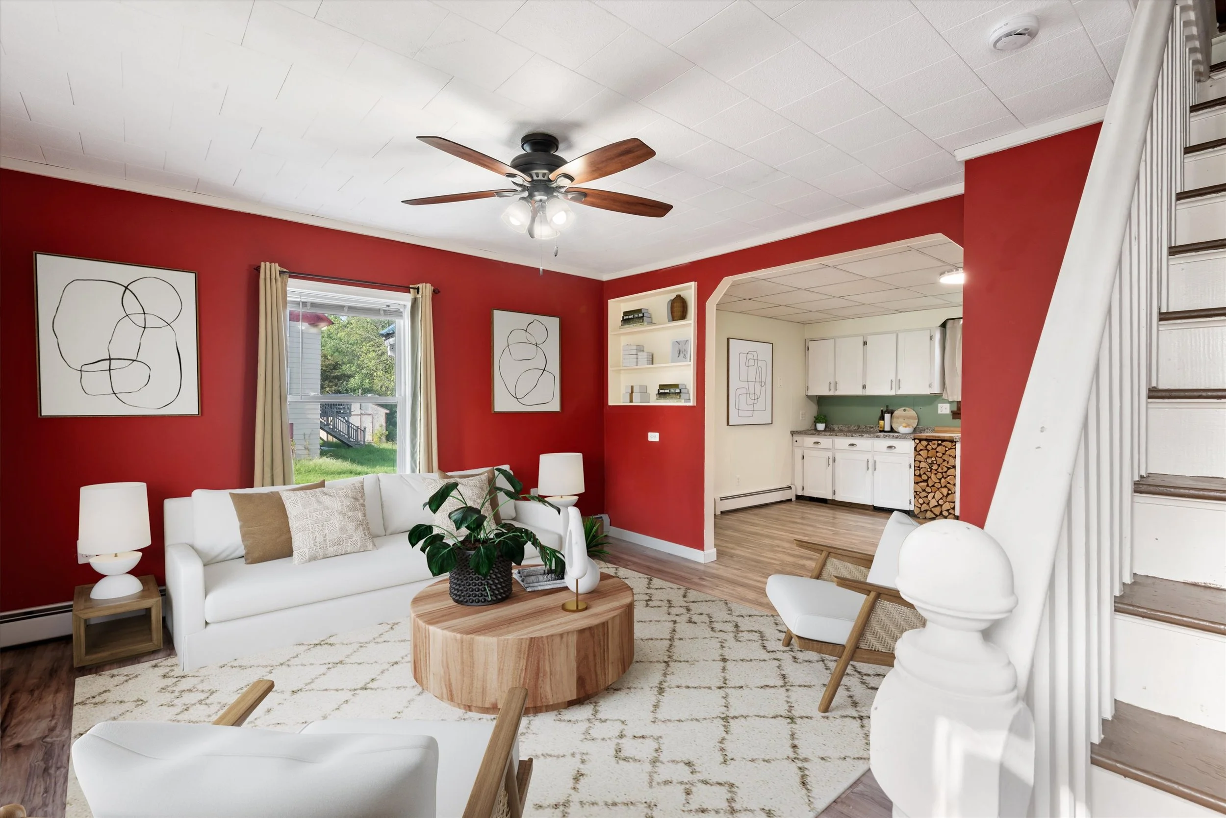Living room with white sofa, beige pillows, white armchair, plants, a round wooden coffee table, and framed artwork on a red wall.