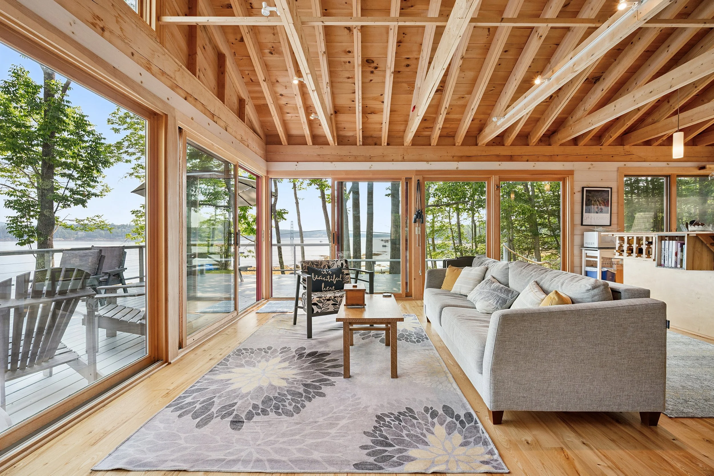 Living room with large glass sliding doors opening to outdoor deck overlooking a lake, wooden high-vaulted ceiling, light-colored sofa, patterned area rug, and outdoor furniture visible through the windows.