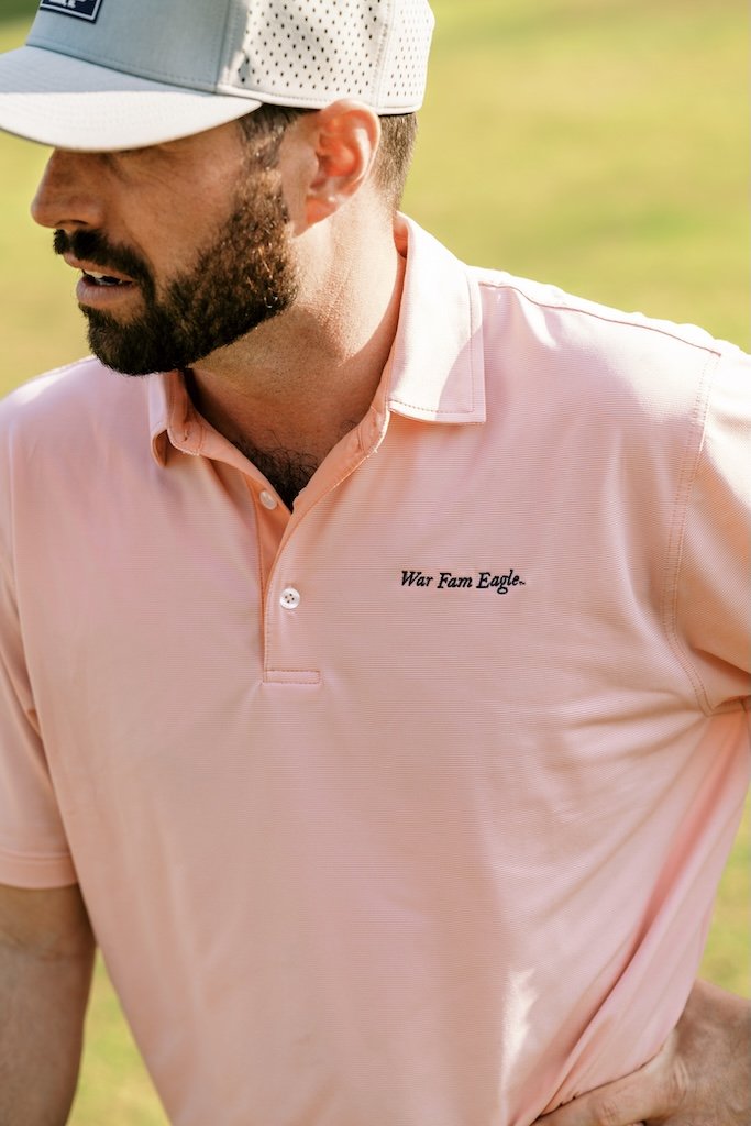 Man on golf course wearing War Fam Eagle's Salmon and White Men's Polo with Script "War Fam Eagle" logo on left chest in navy, and wearing cloud grey War Fam Eagle hydro hat.