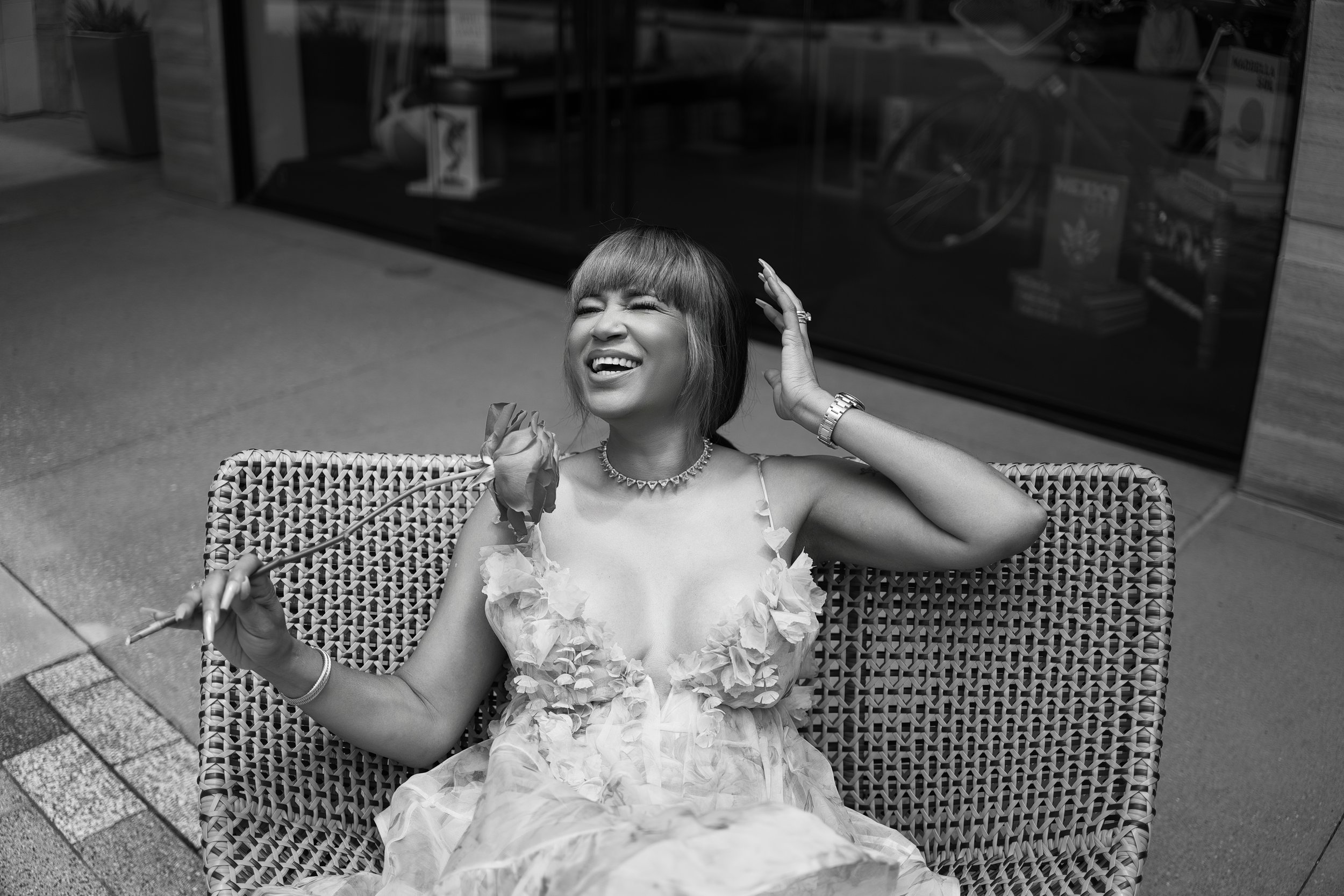 A woman in a floral dress sitting on a wicker bench, laughing, holding a rose, and dressed in jewelry, with a bookstore display in the background.