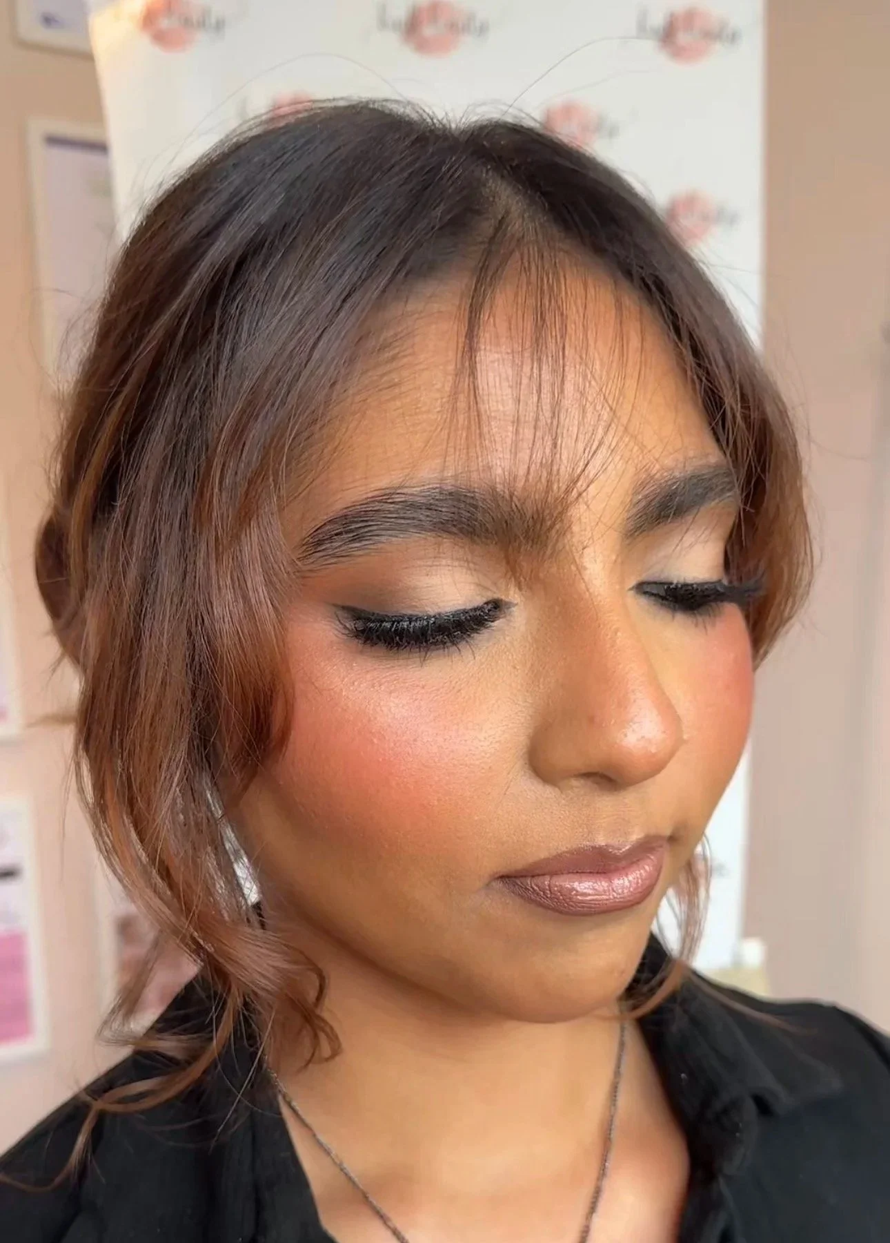 A woman with glam makeup, winged eyeliner and gluffy lashes. Flawless skin with a rosy flushed cheek and a nude glossy lip/