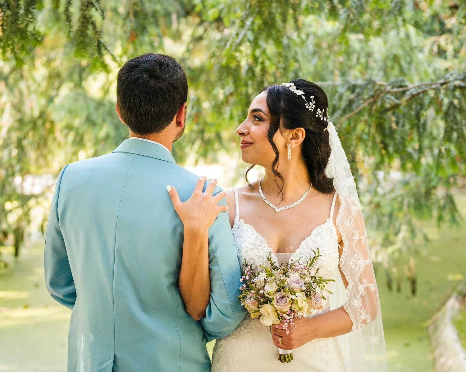 Bruid en bruidegom bij een meer met groene bomen op de achtergrond, trouwjurk, man in lichtblauwe pak, blij in elkaars gezelschap.