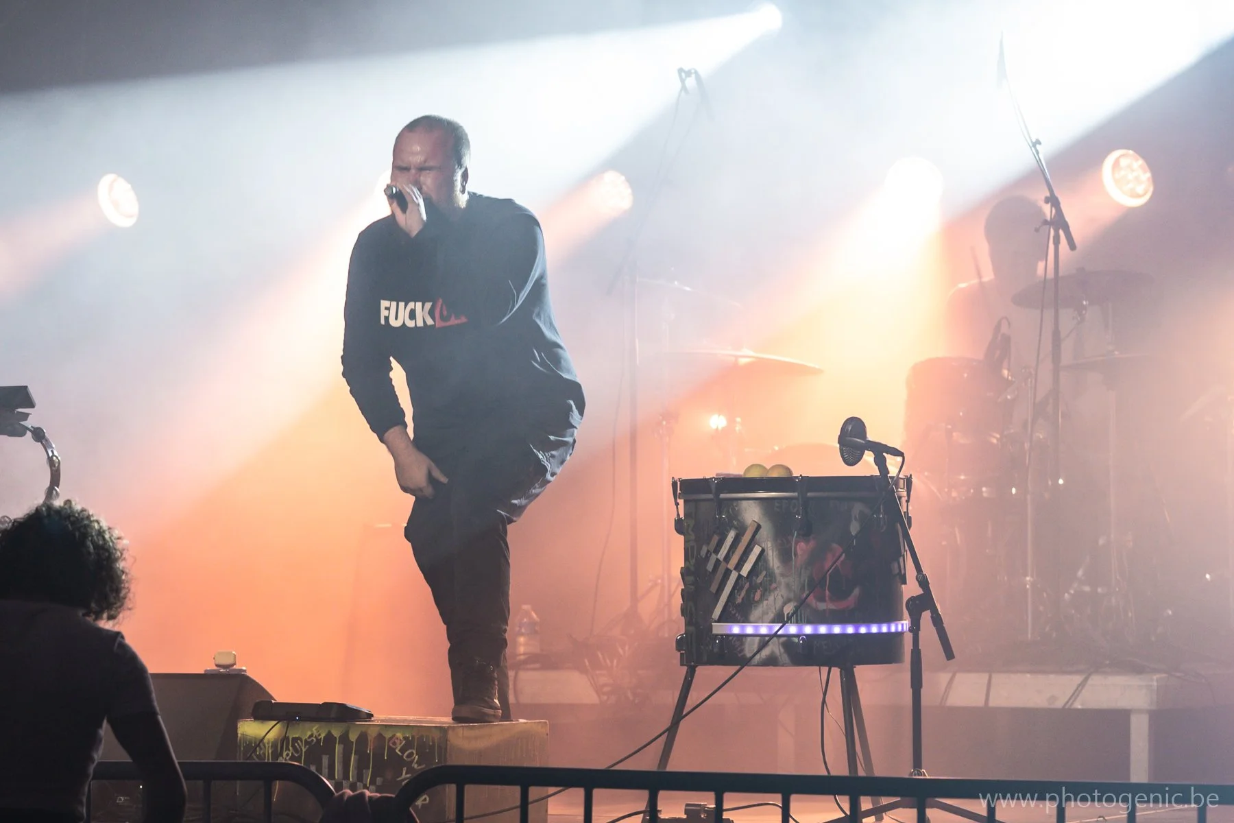 Persoon op podium zingt in microfoon tijdens concert met emotionele uitdrukking, omgeven door lichteffecten en bandinstrumenten.
