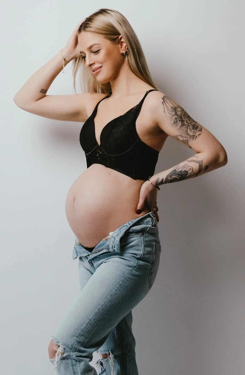 Zwangere vrouw met tattoos draagt zwarte top en blauwe jeans, rust op haar heup tegen een witte achtergrond.