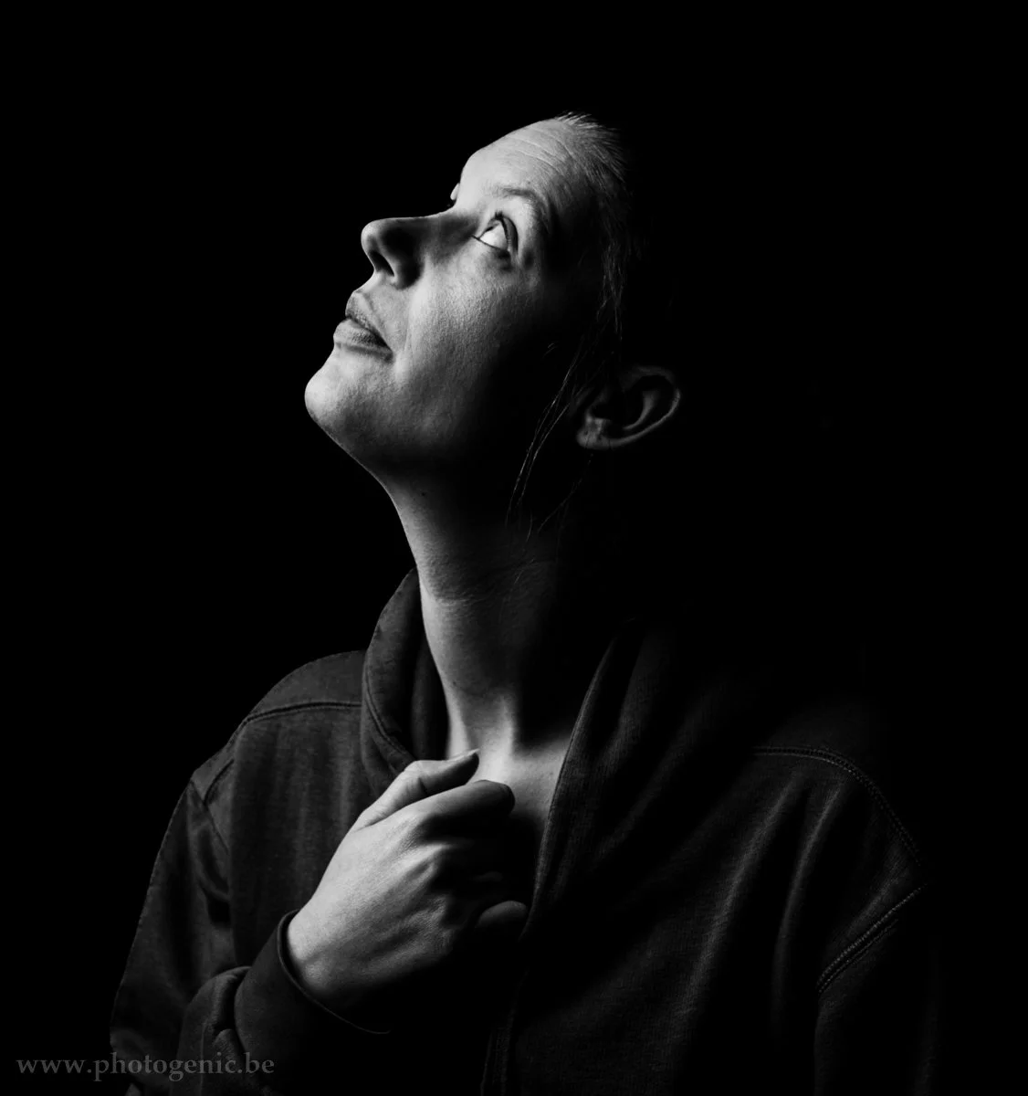 Zwart-witte foto van een persoon die omhoog kijkt, met de hand op de borst en een donkere achtergrond.