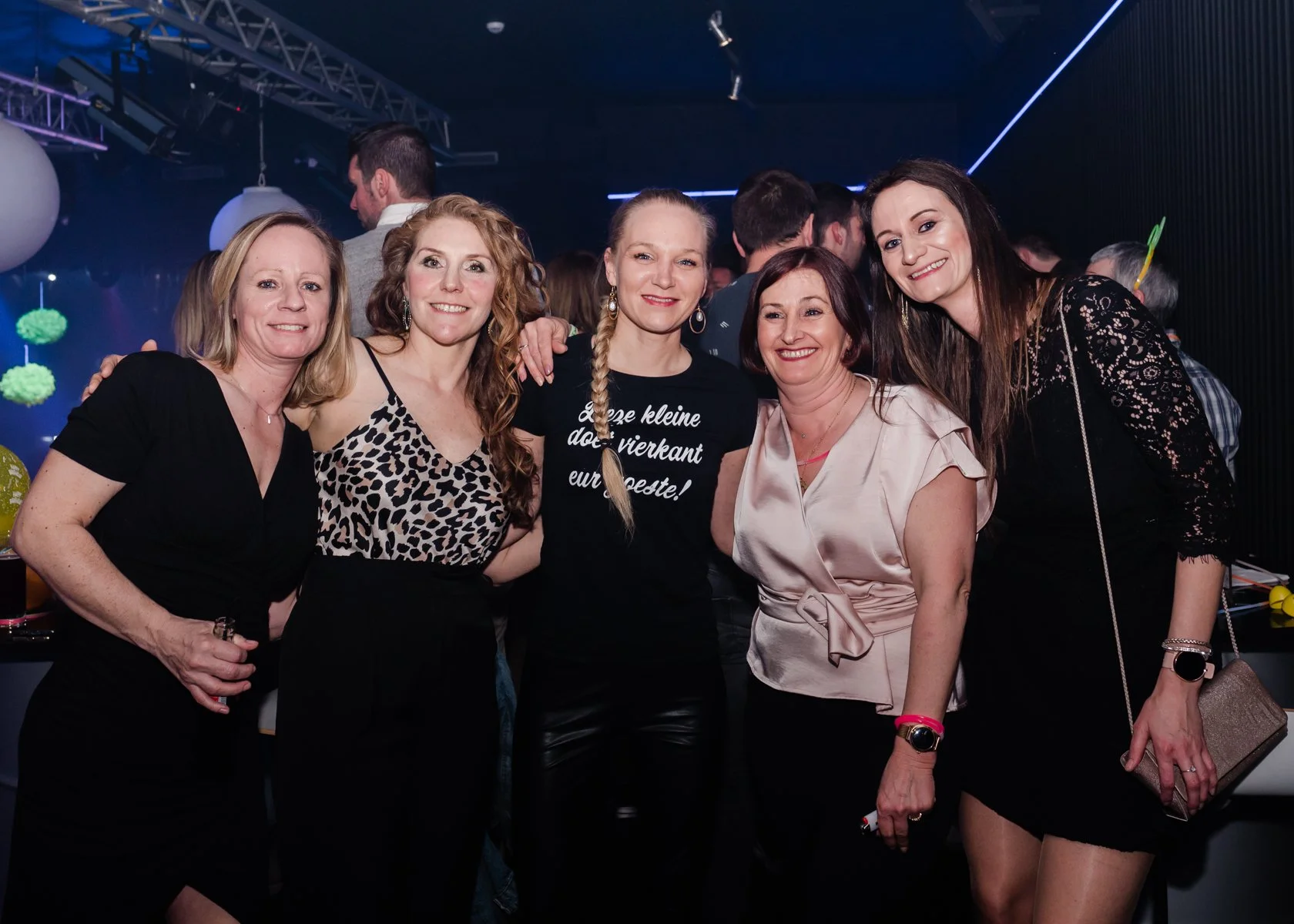 Vijf vrouwen die samen lachend poseren op een feestje of evenement, met een donkere dansvloer en andere feestgangers op de achtergrond.