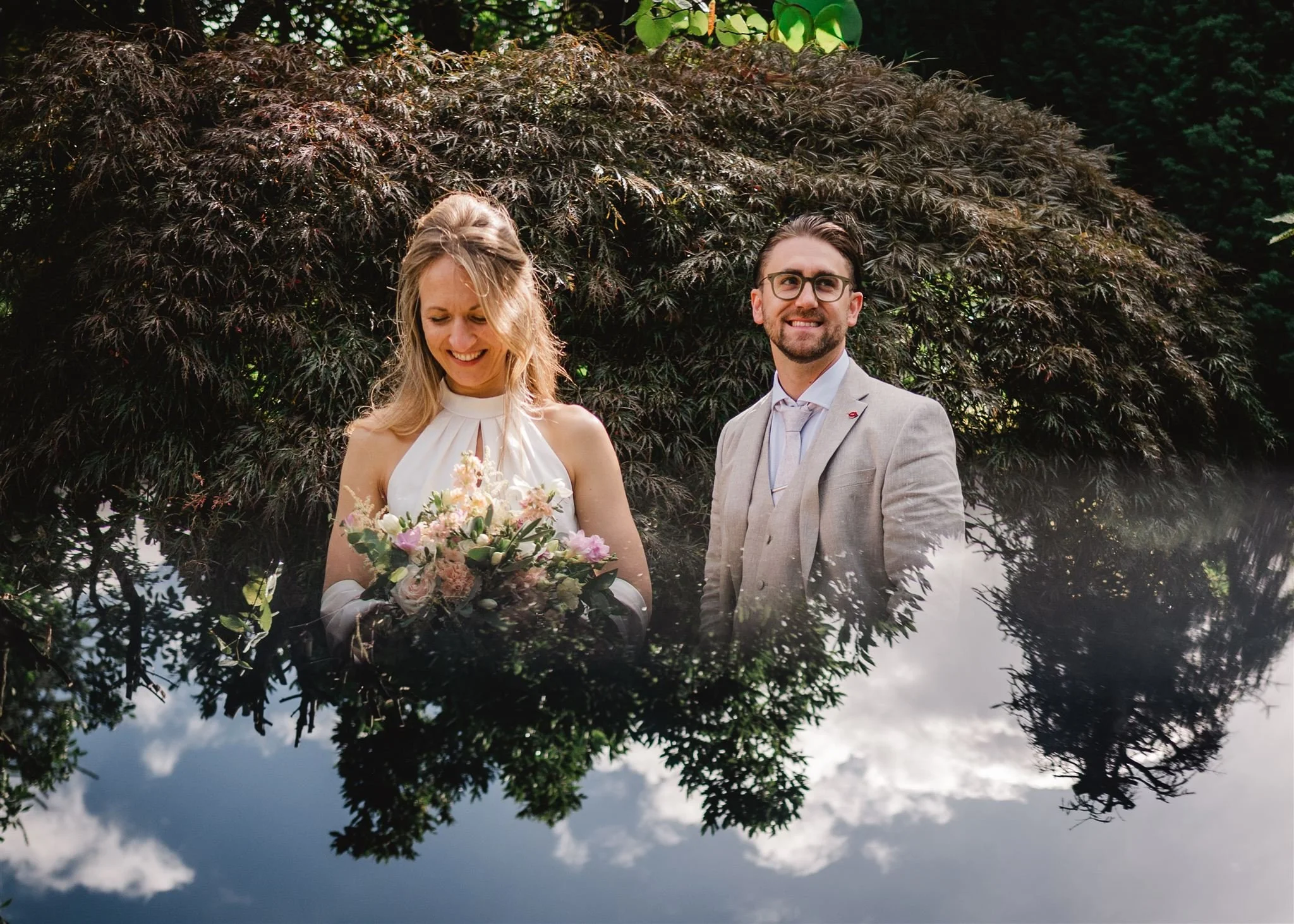 Een getrouwde stel op een photographische spiegeling met een bosachtergrond. De vrouw houdt een boeket bloemen vast en draagt een witte jurk, terwijl de man een lichtgrijze pak met een witte blouse en stropdas draagt. Ze glimlachen en staan naast elk