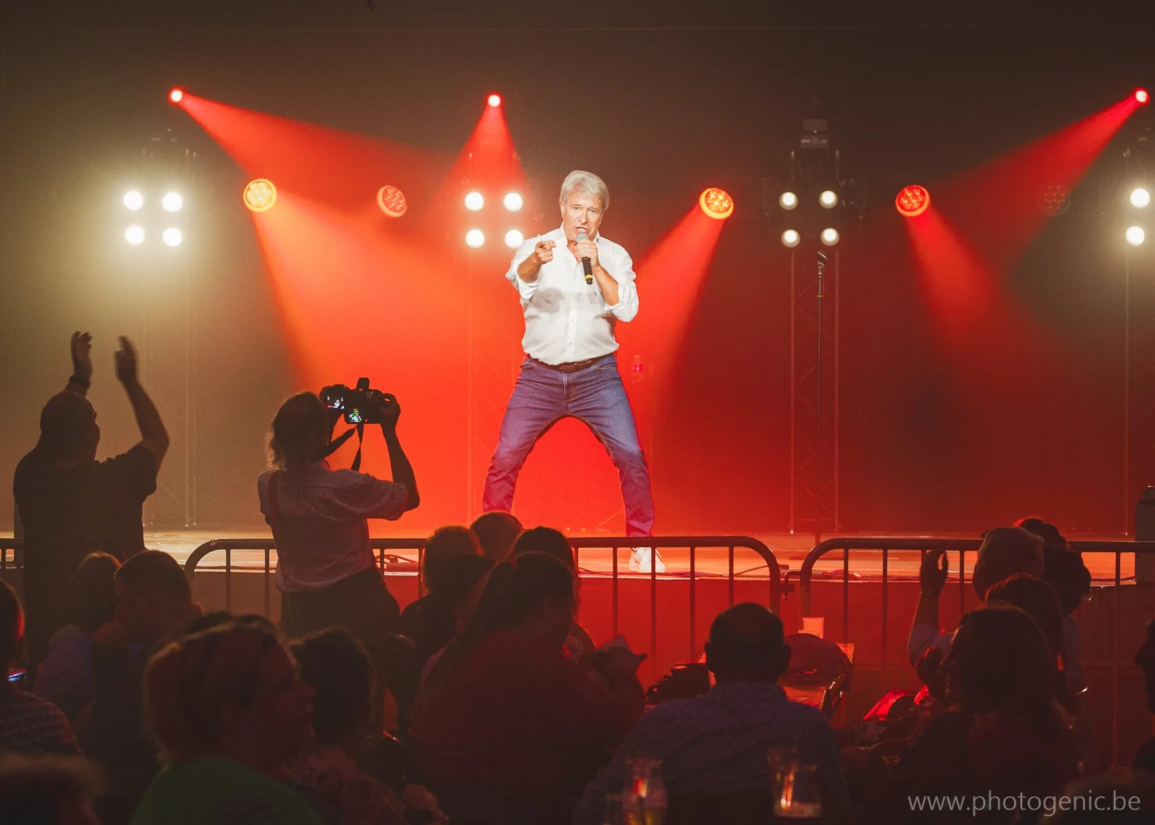 Man op een podium tijdens een optreden, met rode lichtstralen op de achtergrond, terwijl een publiek kijkt en een persoon met een camera opneemt.