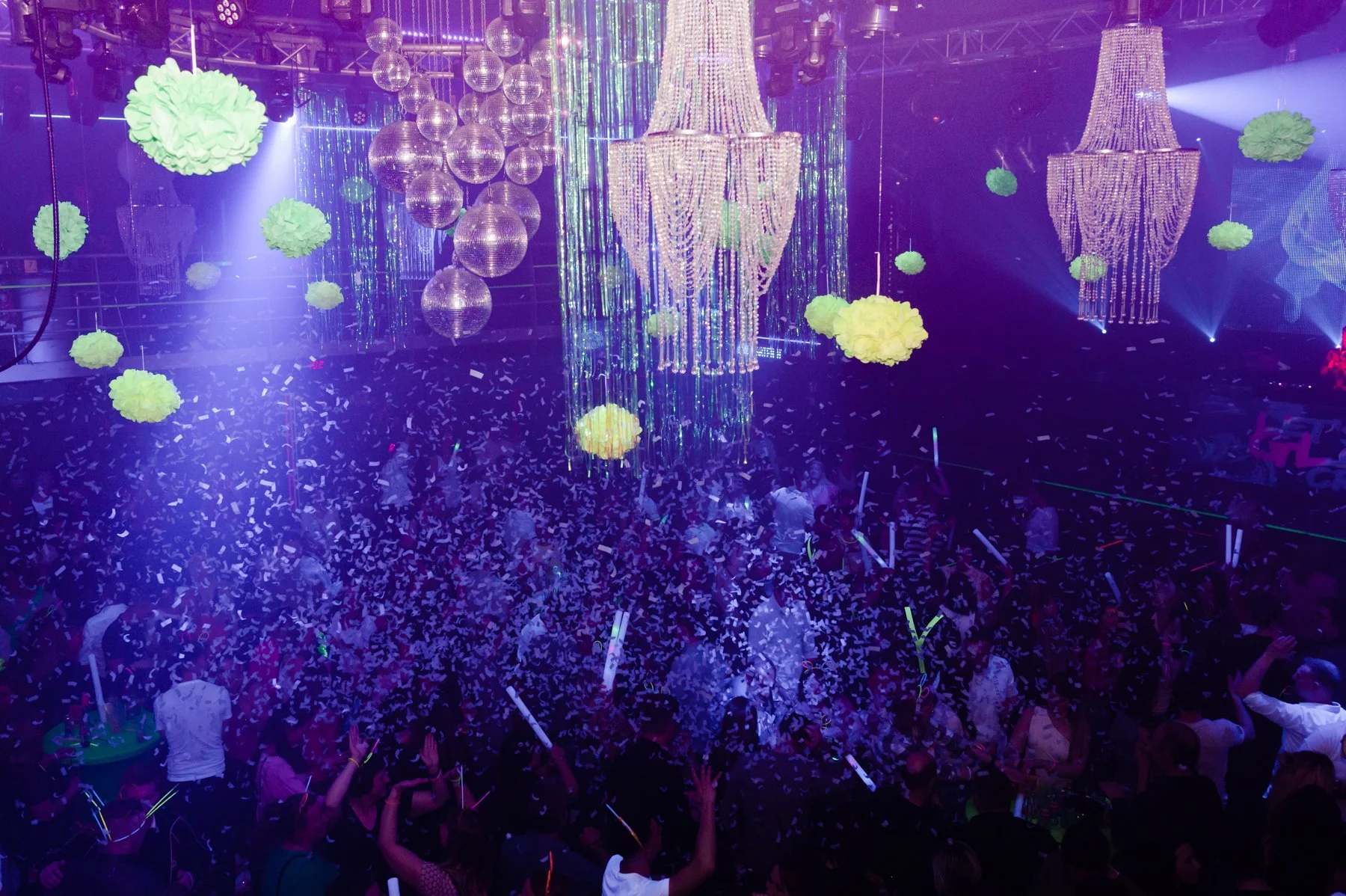 Feestzaal met grote kroonluchters, kleurrijke verlichting, groene pompon decoraties, en een dansende menigte met confetti en glowsticks.