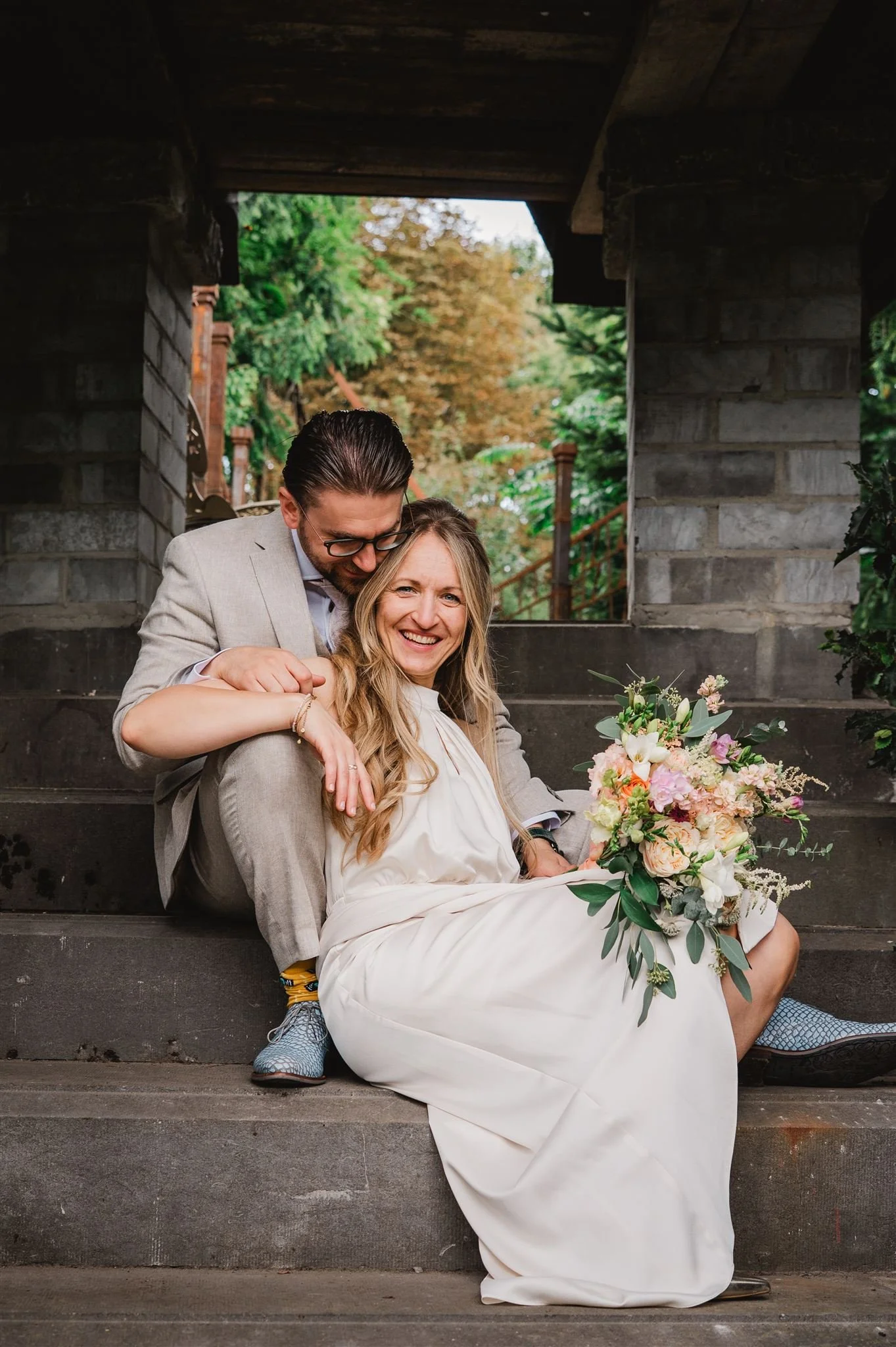 Een gelukkige vrouw in een witte jurk zit op de trappen met een grote bloemenstronk, terwijl een man in een lichtgrijs pak naast haar ligt en zich over haar buigt.