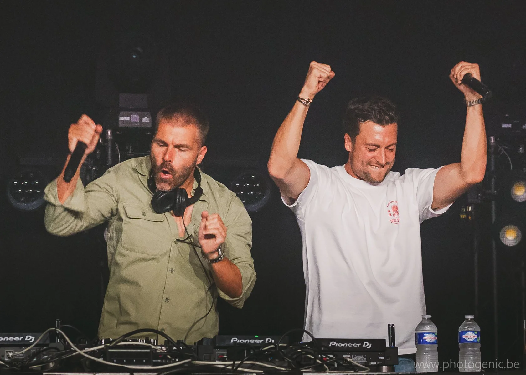 Twee mannen achter DJ-sets, de man aan de linkerkant houdt een microfoon en danst met ogen gesloten, de man aan de rechterkant viert trots met opgewonden handen omhoog, beide op een donkere achtergrond met DJ-apparatuur.