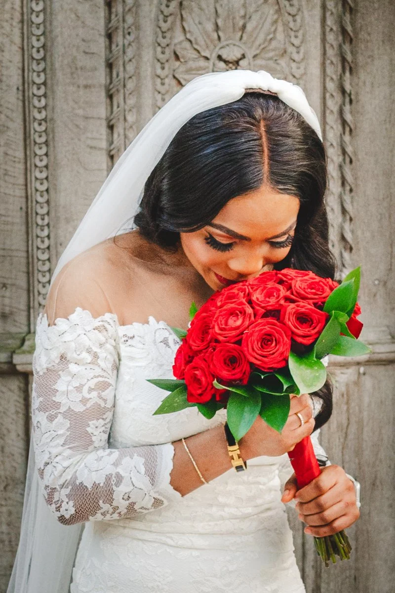 Een bruid in een witte kanten jurk met een witte sluier die naar een bouquet van rode rozen en groene bladeren kijkt terwijl ze haar hoofd buigt.