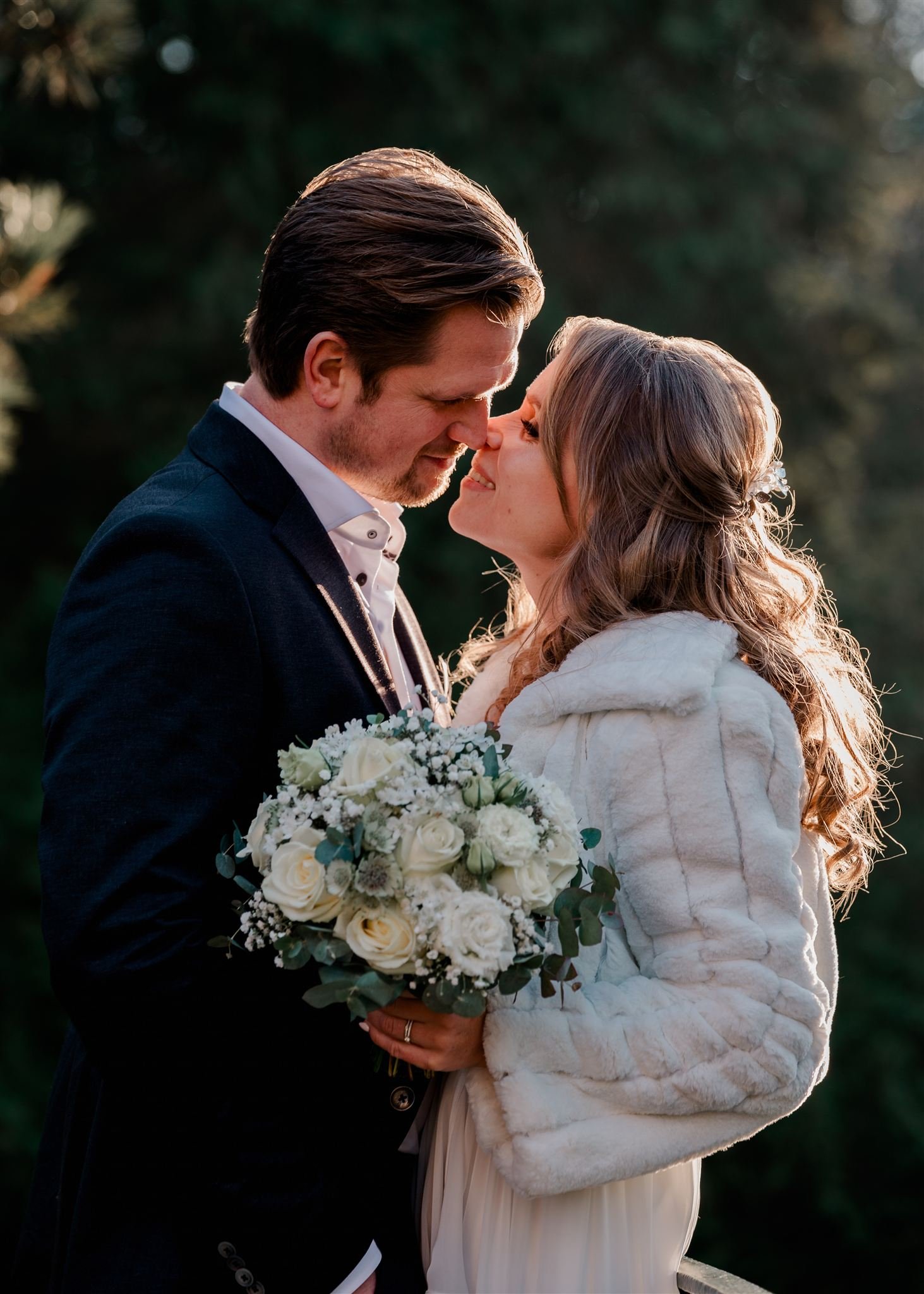 Een man en een vrouw staan dicht bij elkaar, kijken elkaar liefdevol aan, met hun neuzen bijna tegen elkaar, tijdens een buitenceremonie. De vrouw houdt een witte bloemboeket vast en draagt een witte jas. De man draagt een donker pak en een witte shirt.