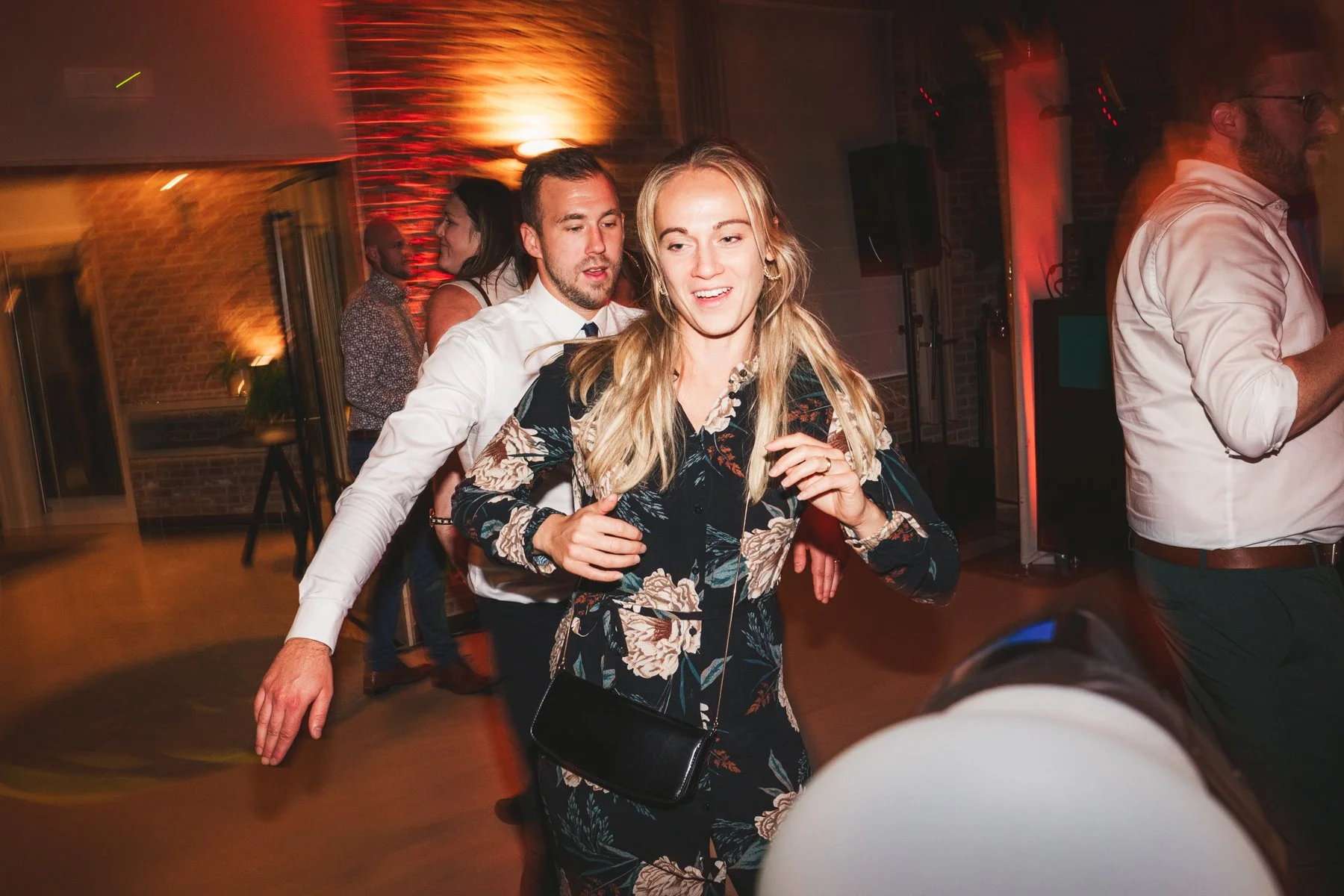 Mensen dansen op een feestje in een zaal met bruine bakstenen muren, met kleurrijk en dynamisch licht.