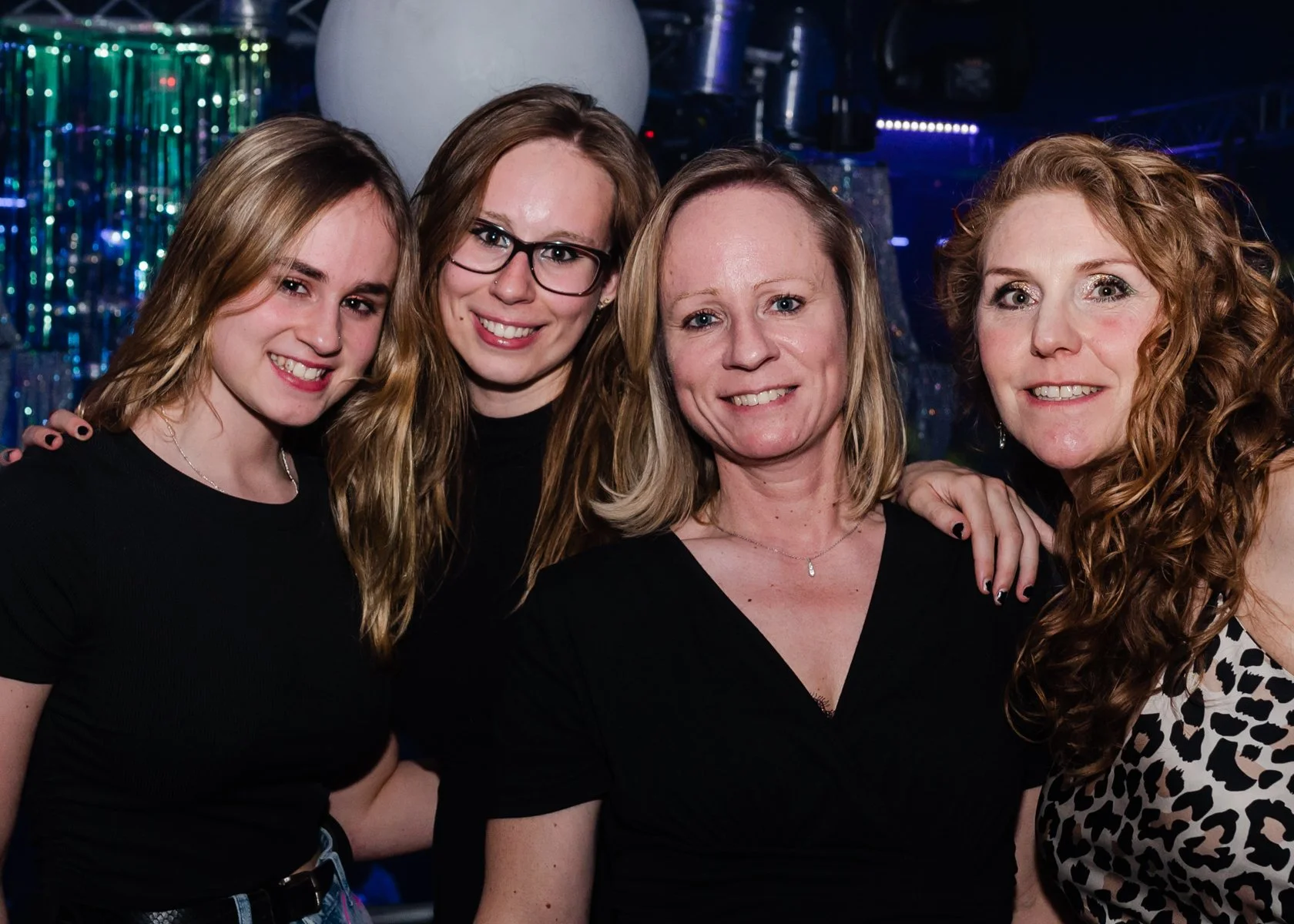 Vijf vrouwen poseren samen op een feest met kleurrijke decoraties op de achtergrond.