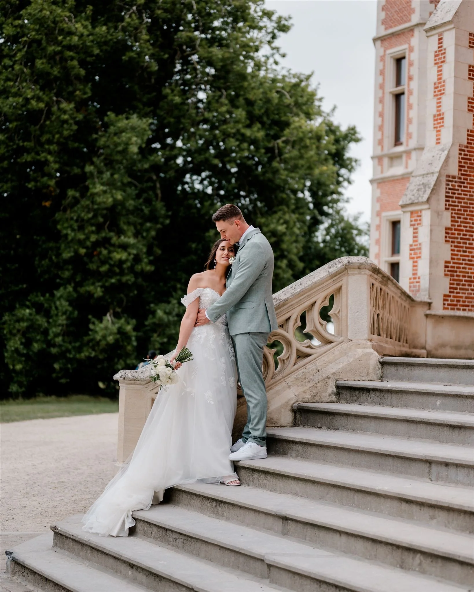 Een bruid en bruidegom staan samen op de trappen van een kasteel, de bruid houdt een boeket witte bloemen vast, ze lijken te genieten op hun huwelijksdag.