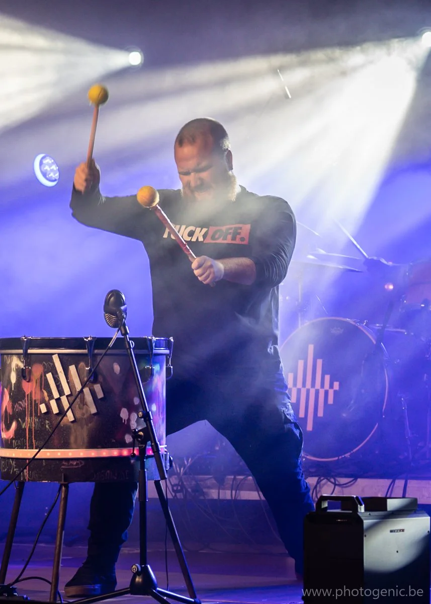 Man speelt de drums met twee hamerachtige stokken, op een podium met kleurrijke lichten en rook.