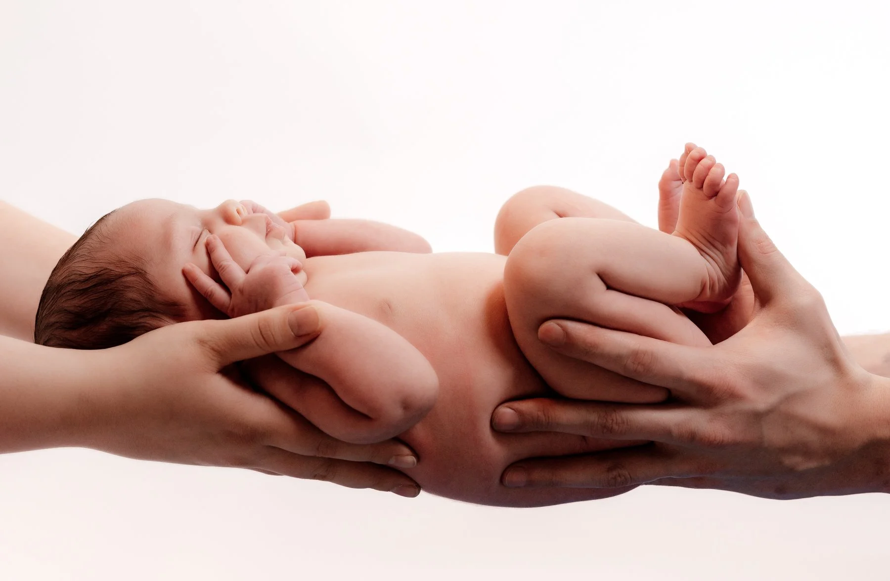 Een pasgeboren baby wordt vastgehouden door meerdere handen, met een witte achtergrond.