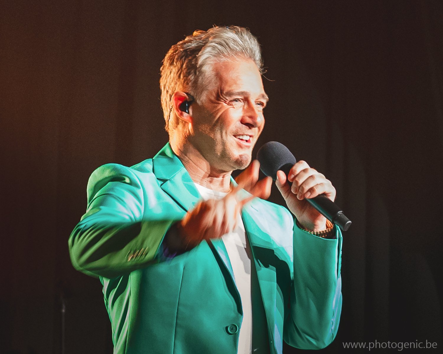 Een man in een groene jas staat op een podium en houdt een microfoon vast, lachend en gebarend met zijn handen.