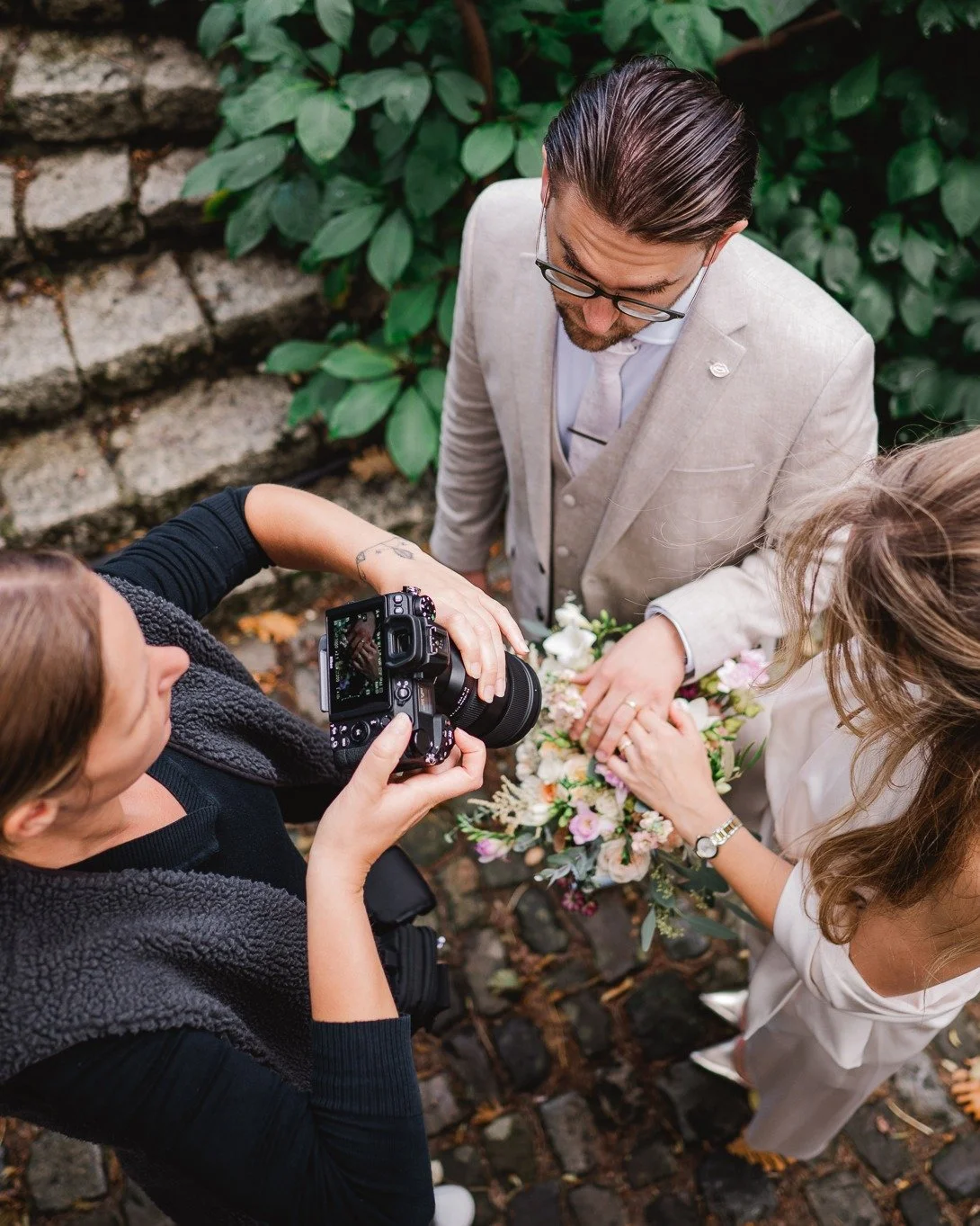 Een groep mensen die een huwelijksceremonie houdt in een tuin, met een bruid en een bruidegom die hand in hand staan, terwijl een fotograaf foto's maakt.