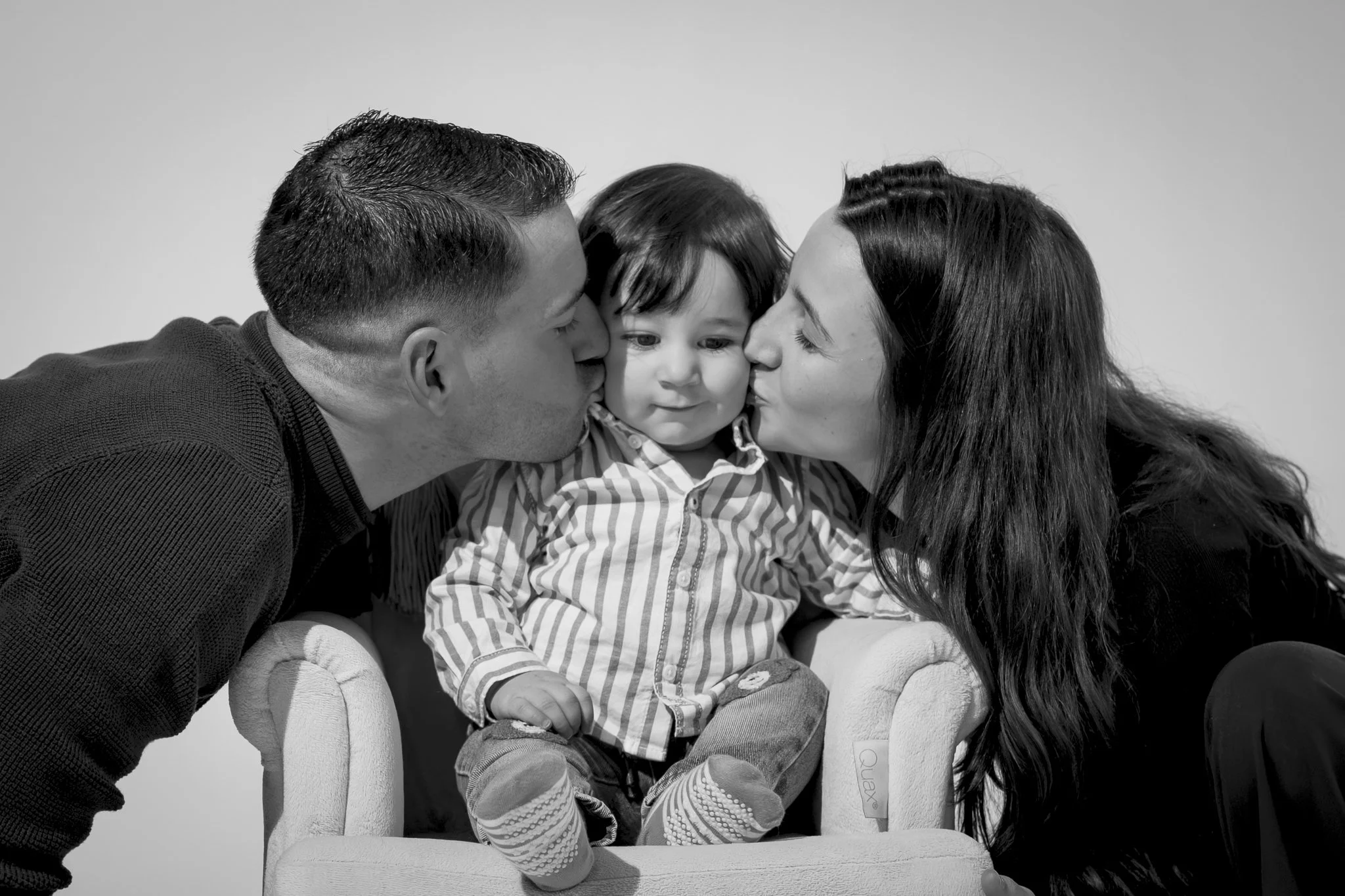 Twee volwassenen kussen een jongen op iedere wang, terwijl de jongen op een kinderstoel zit, in zwart-wit.