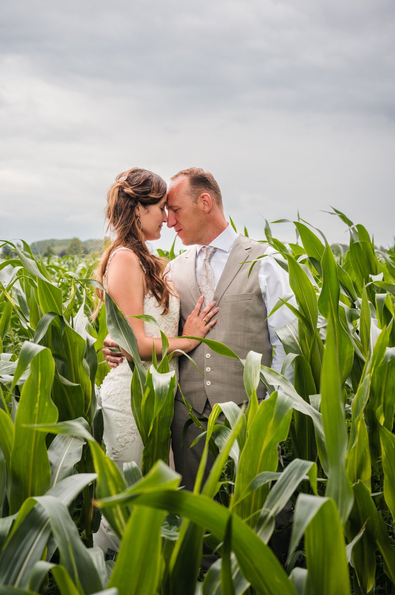 Een getrouwd koppel in formele kleding, omringd door een veld met gekweekte maïsplanten, met een bewolkte hemel op de achtergrond.