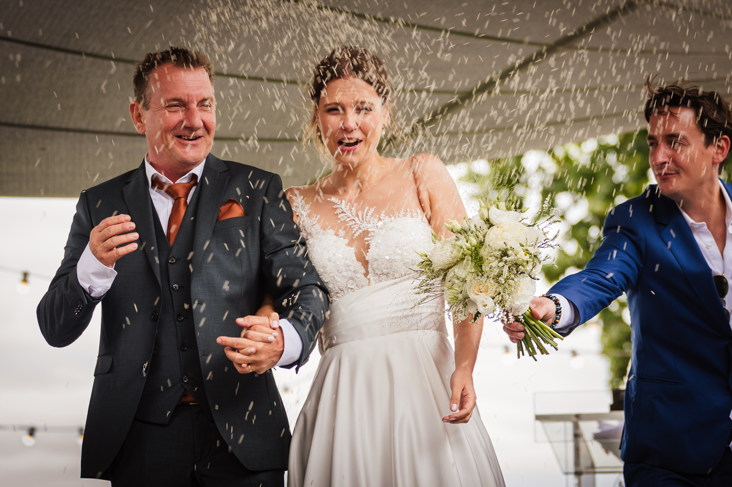 Bruidegom en twee mannen in pakken, een vrouw in een witte trouwjurk met bloemen, feestvierend met confetti tijdens een bruiloft.