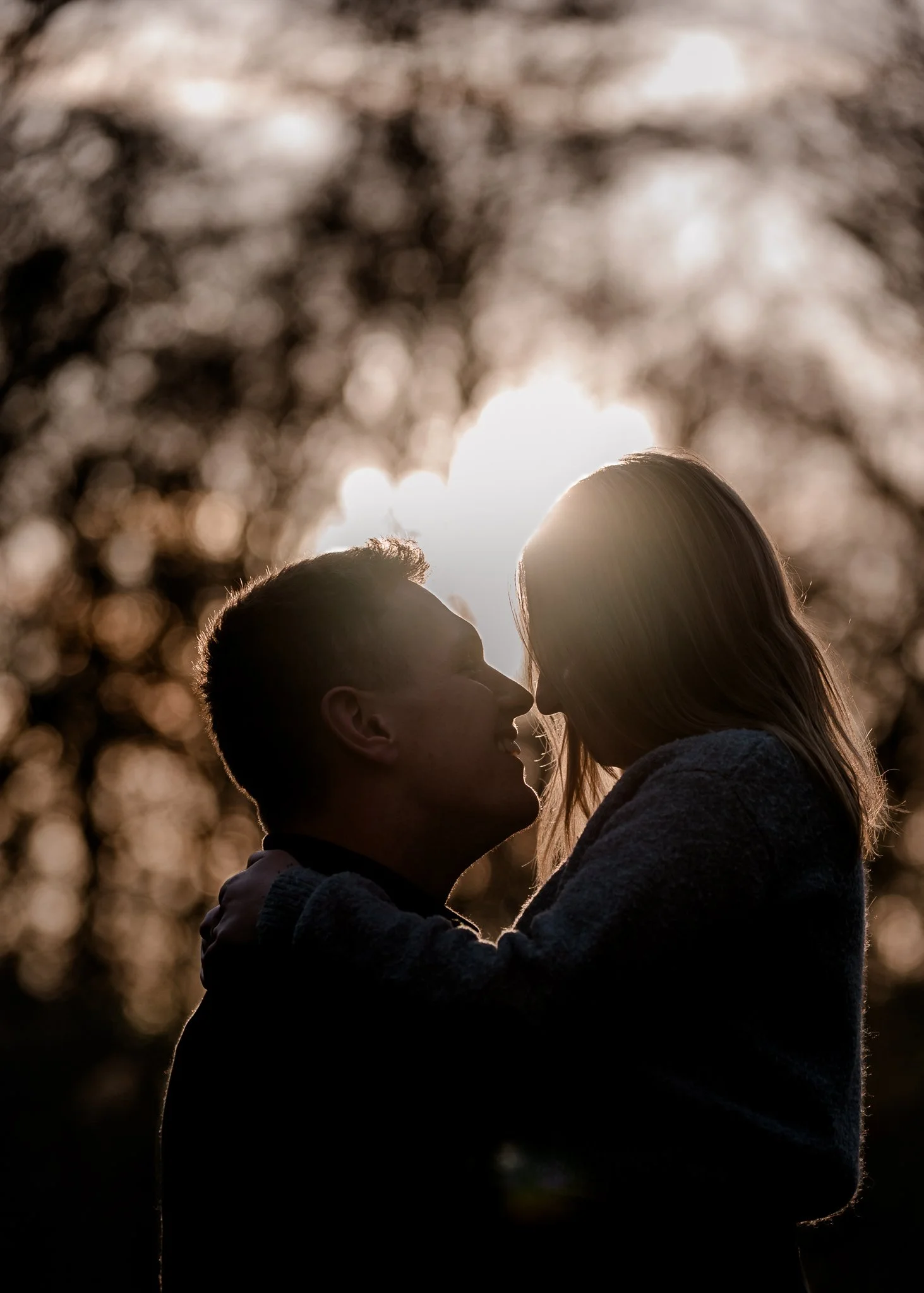 Silhouet van een man en een vrouw die dicht bij elkaar staan, met de zon op de achtergrond en bladeren in de omgeving.