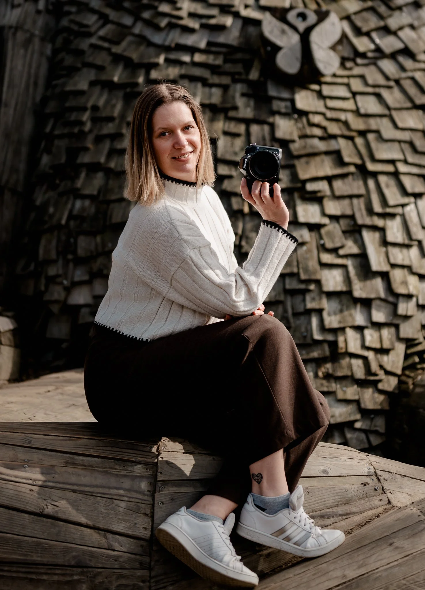 Een vrouw zit op een houten bankje, houdt een camera in haar rechterhand en posereert voor een achtergrond van houtschijven en een vleugelvormige decoratie.