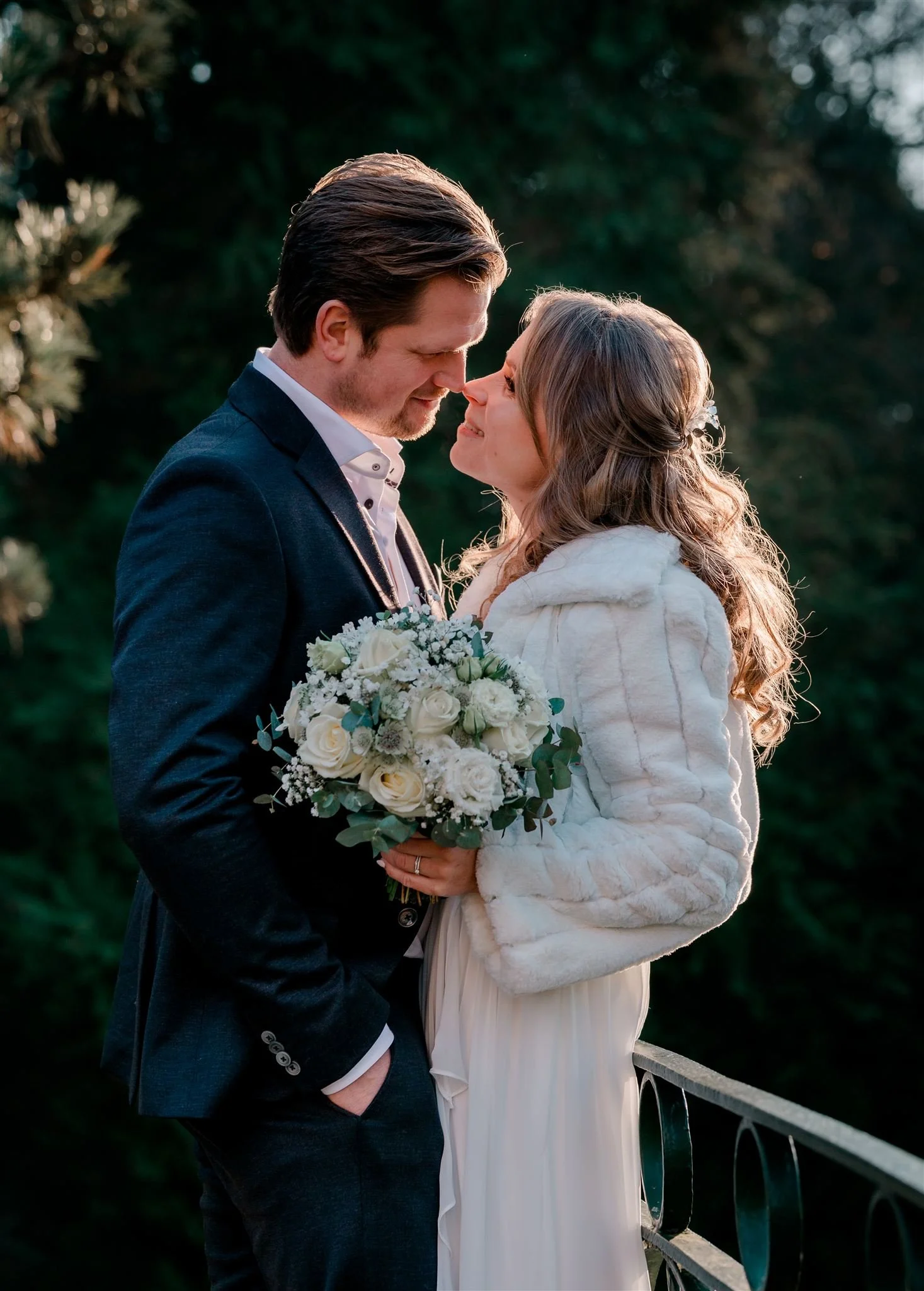 Een bruiloftsfoto van een koppel die dicht bij elkaar staan, met hun gezichten bijna aanraken. De vrouw houdt een boeket witte bloemen vast en draagt een witte jurk en jas, terwijl de man een donker pak en witte overhemd draagt. Ze staan buiten tegen