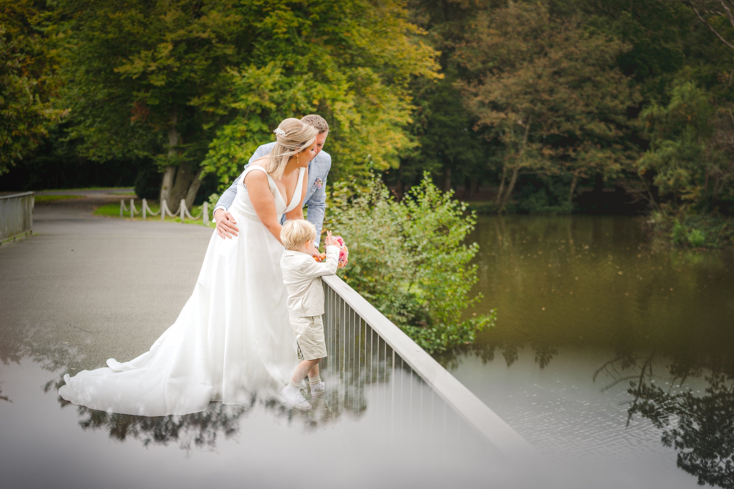 Bruiloftspaar met een kind kijkt over een waterkant vanaf een brug in een park, omringd door bomen.