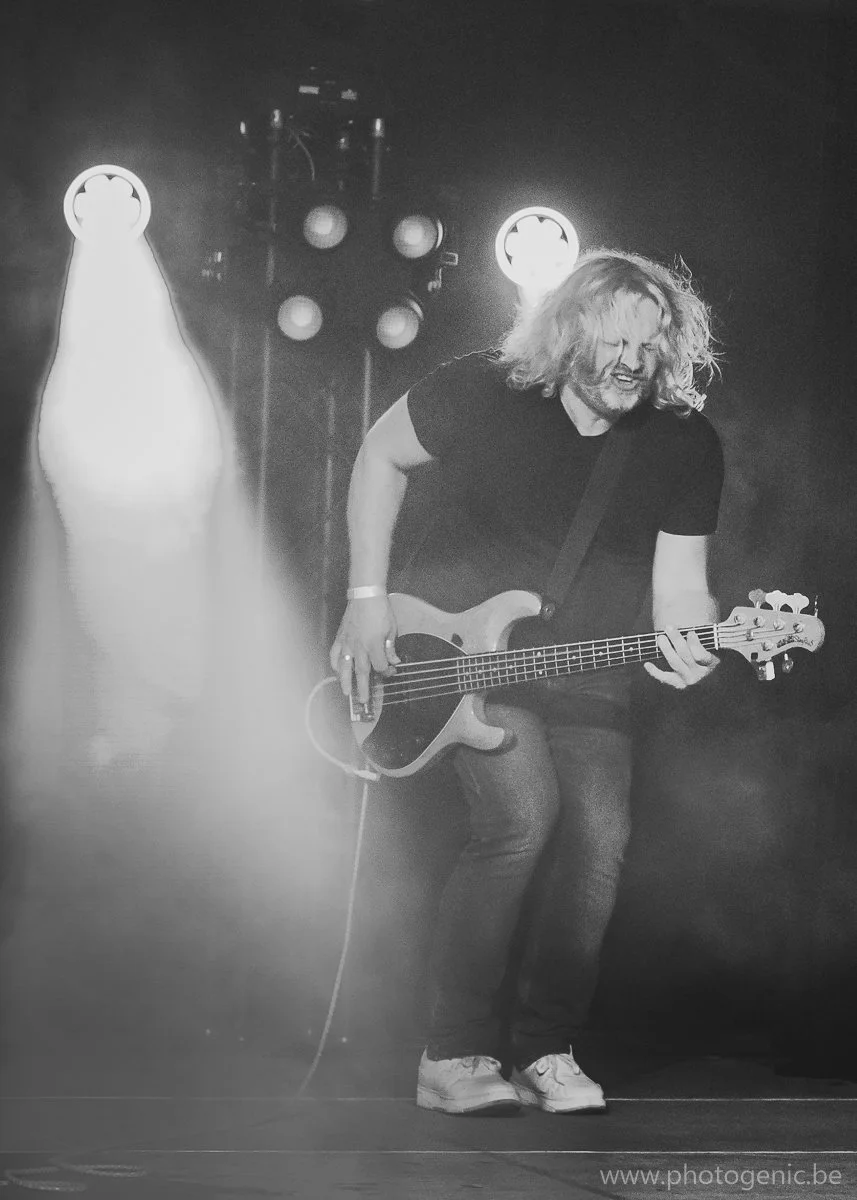 Mannelijke muzikant met lang haar speelt basgitaar op een podium, omringd door podiumverlichting, in zwart-wit.