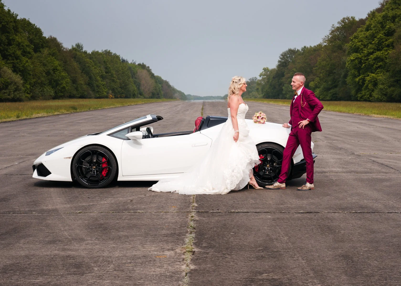 Een bruid in een witte trouwjurk en een bruidegom in een bordeaux kleurige pak staan naast een witte sportwagen op een verlaten weg, met een bos op de achtergrond.