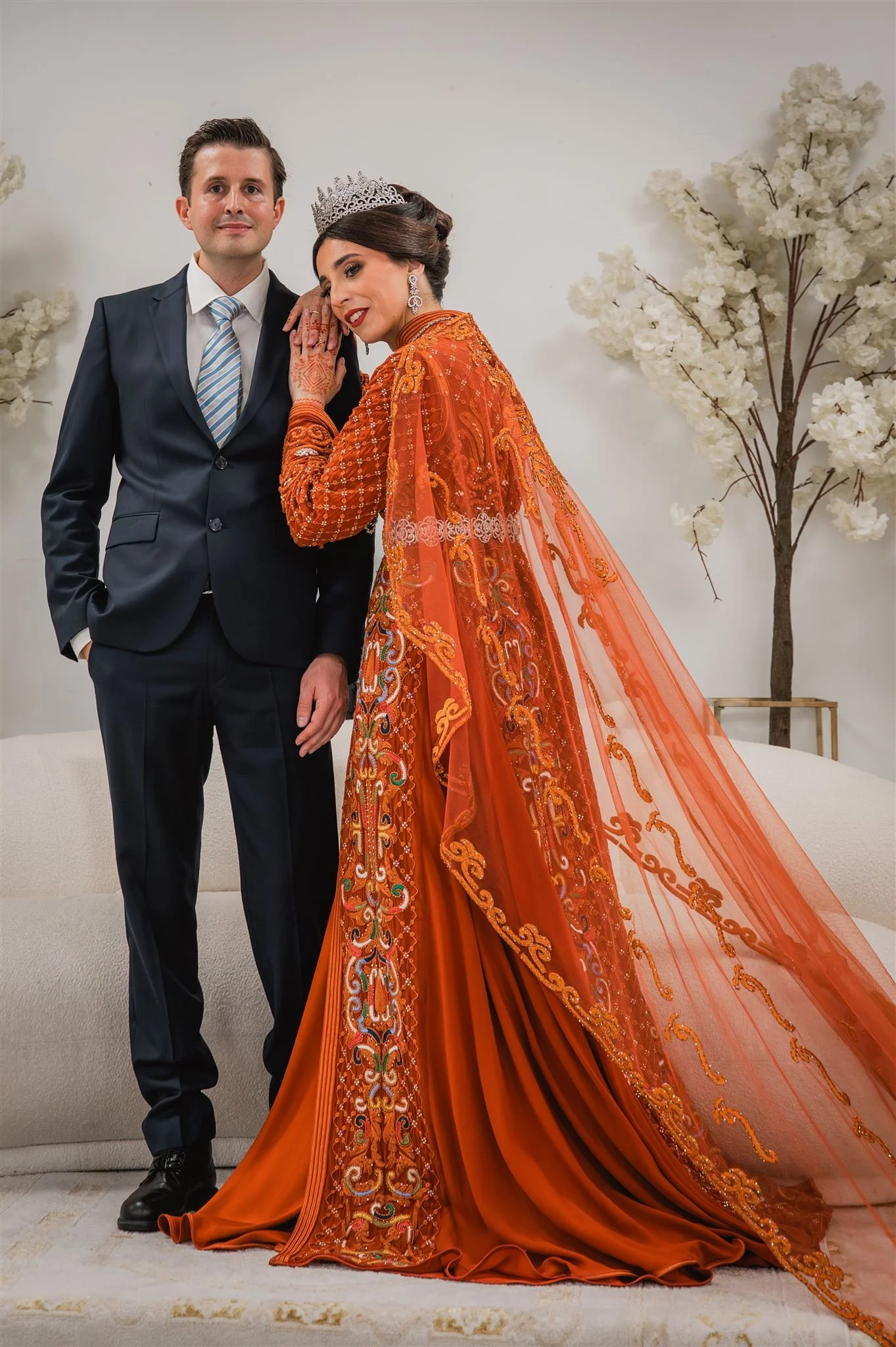 Een man en een vrouw staan samen voor een witte muur. De man draagt een donkerblauw pak met een lichtblauwe stropdas, en de vrouw draagt een oranje, met ingewikkeld borduursel versierde traditionele jurk en een diadeem.