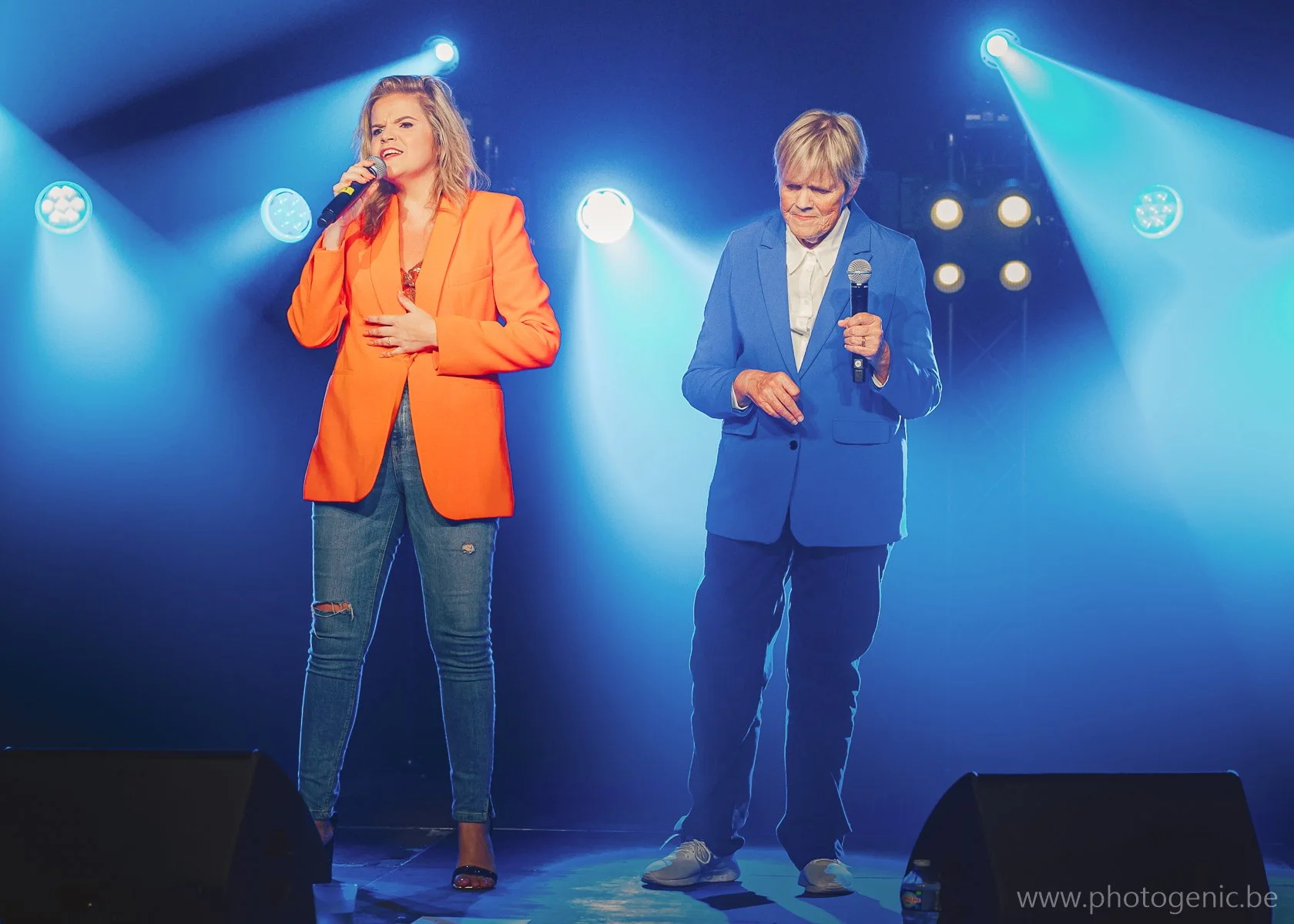 Twee mensen staan op een podium met blauwe en paarse lichten, een vrouw in een oranje blazer en ripped jeans zingt met een microfoon en een oudere man in een blauwe jas en witte schoenen met een microfoon.