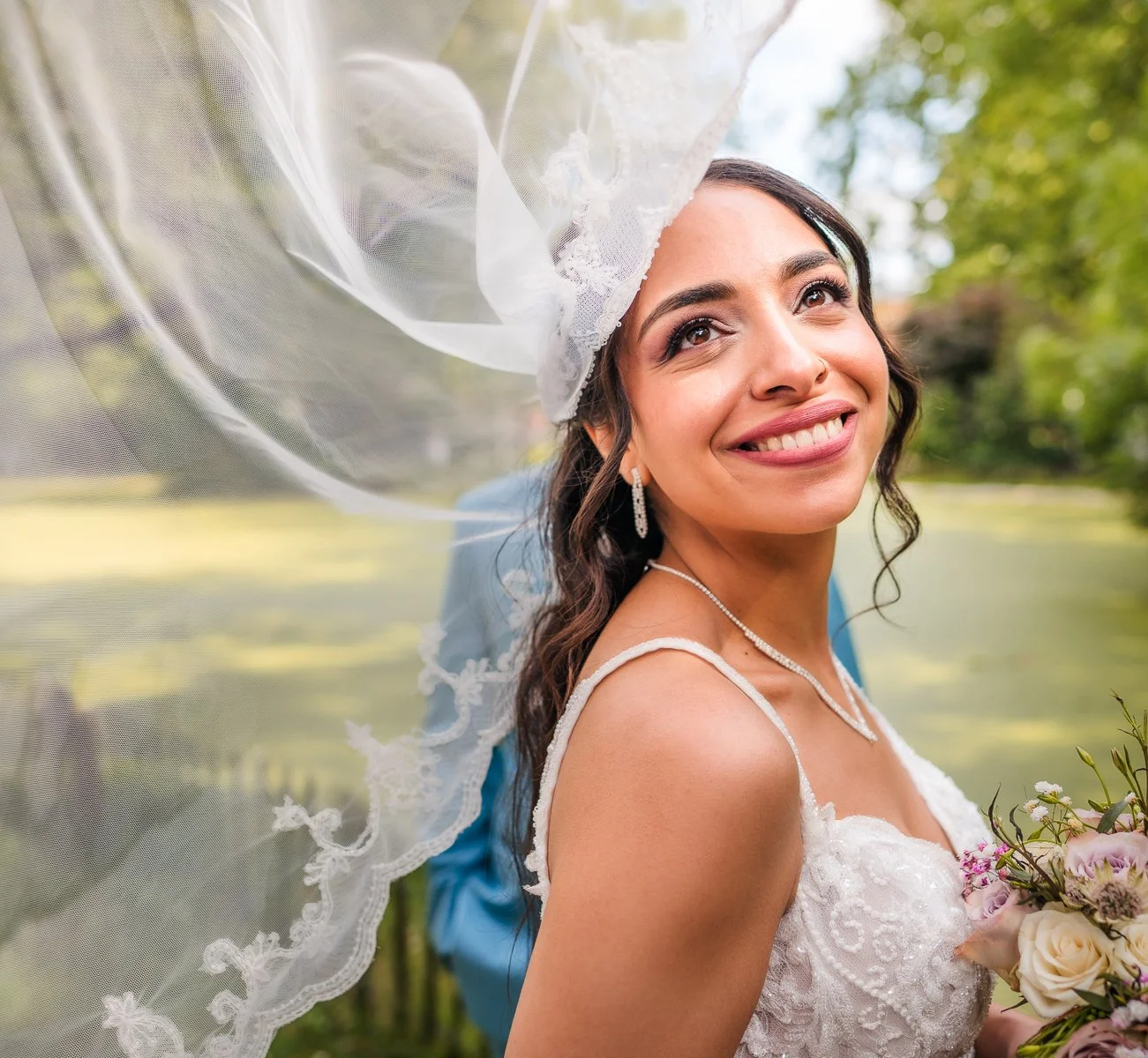 Een glimlachende bruid in een kanten trouwjurk, met een lange voile en sieraden, die buiten in een groene omgeving staat.